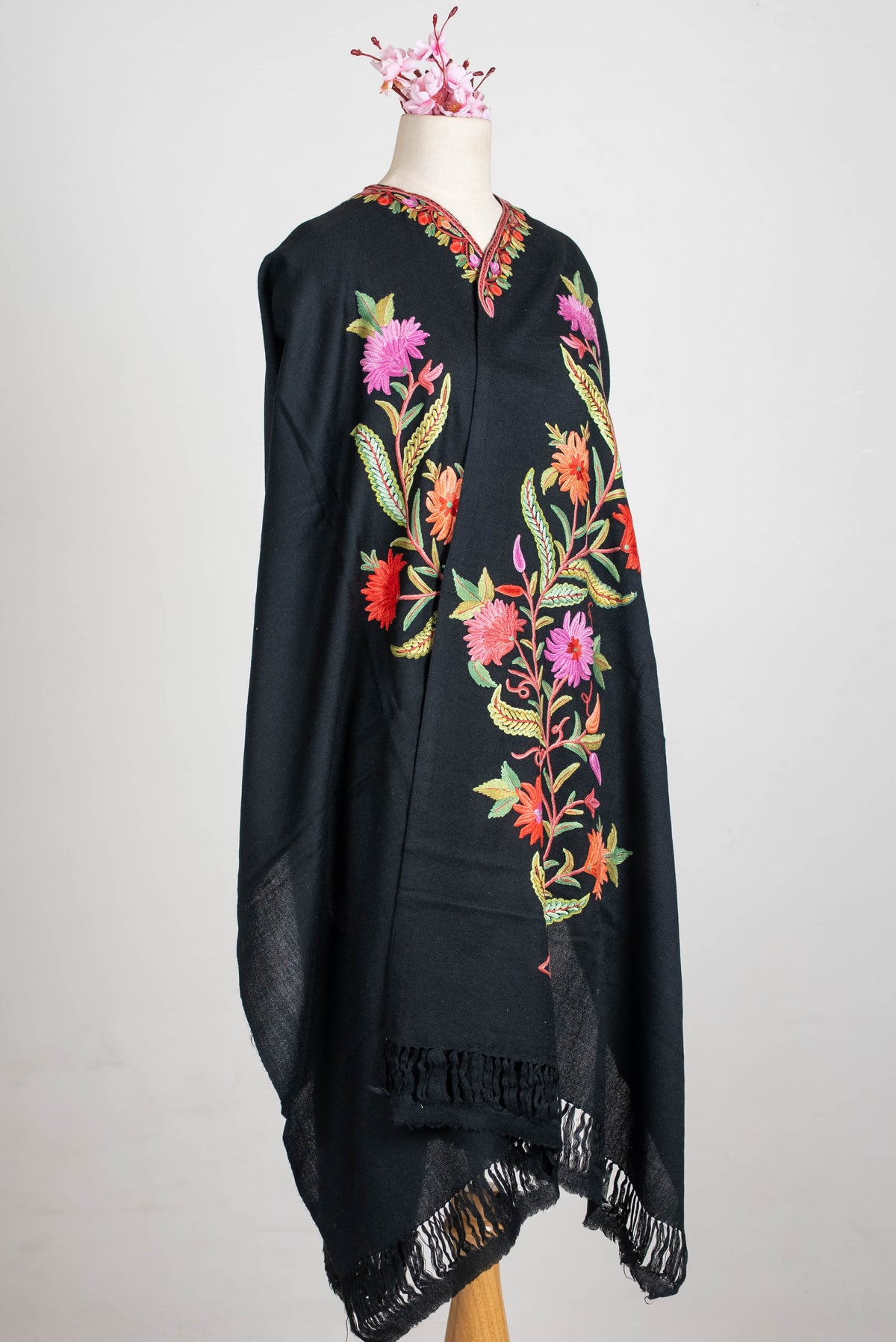 Angled full view of the black shawl cape on a mannequin, showing tall colourful floral stems and fringed ends