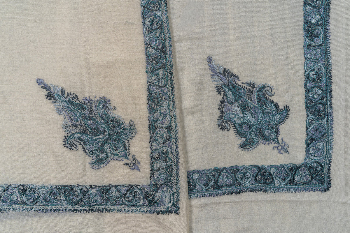 Folded ivory rumal showing the front and reverse of its teal embroidered borders and paisley corner motifs