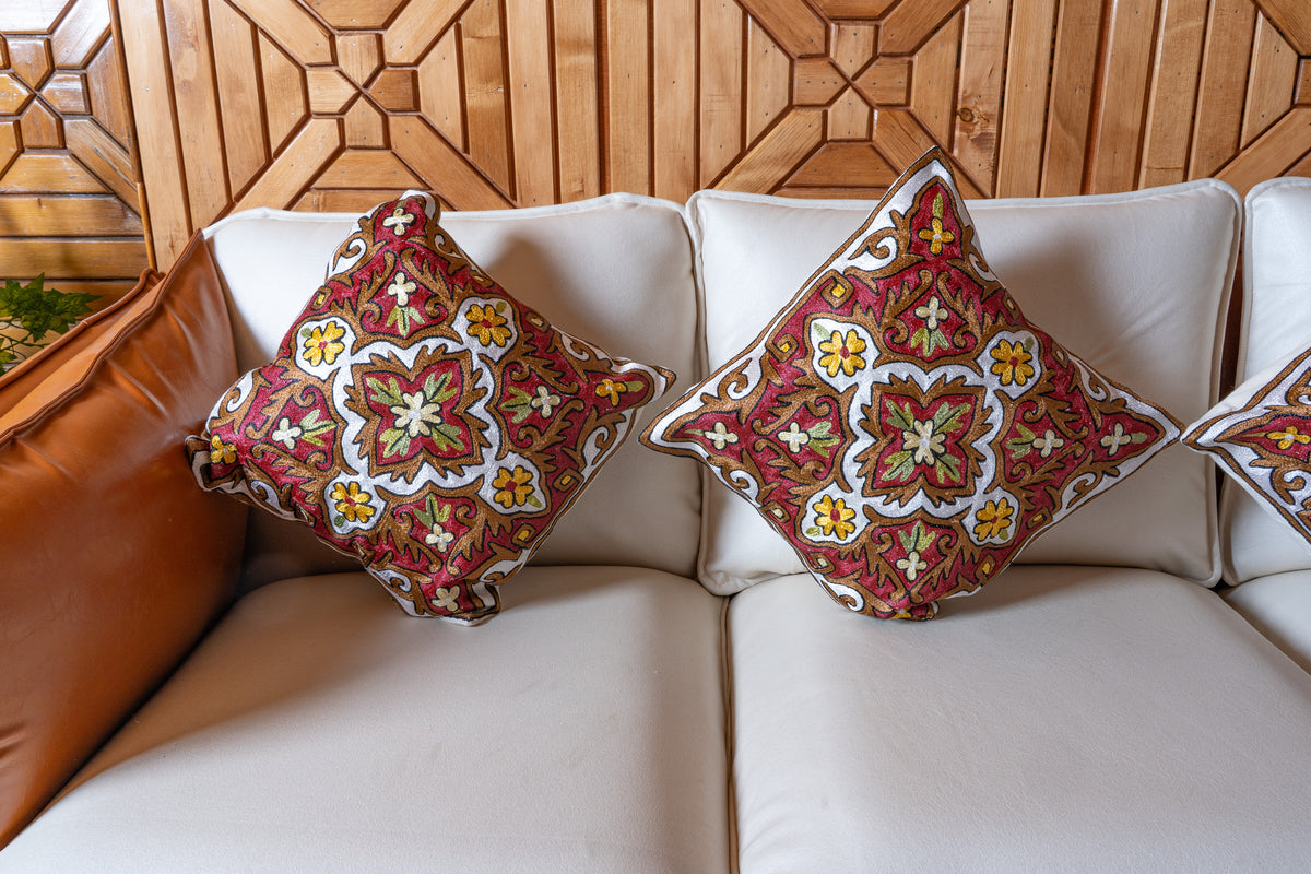 Two matching floral medallion cushions side by side on a cream sofa, with red, ivory, green and yellow embroidery