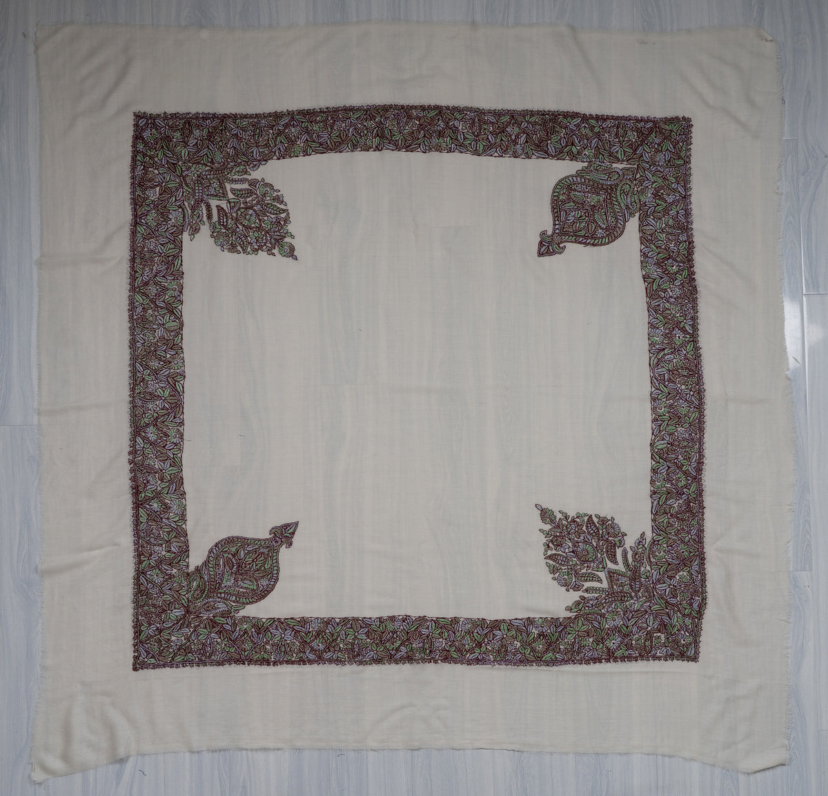 Square ivory Merino wool rumal laid flat, with a purple-and-green floral border and four ornate corner motifs