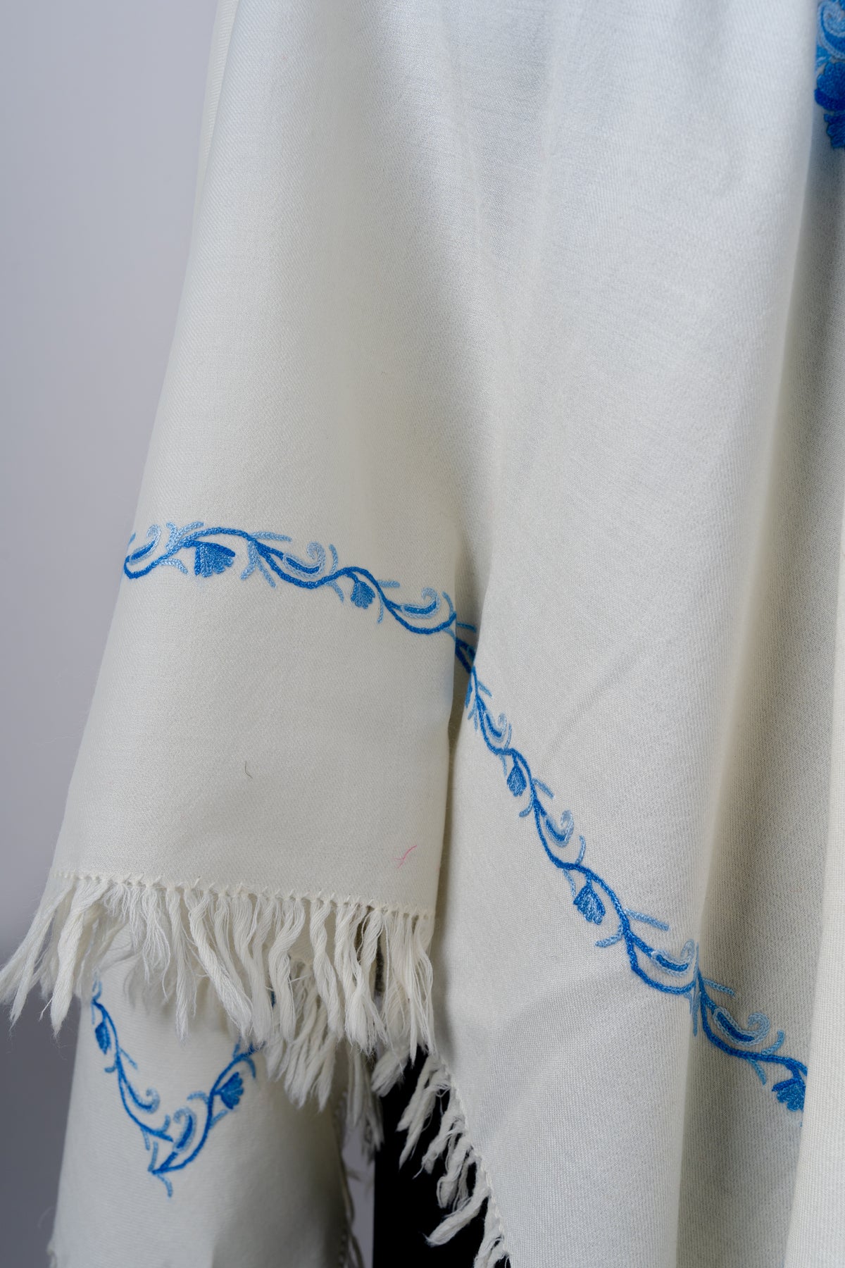 Detail of the ivory poncho side showing a delicate blue curling vine border above soft ivory fringe.
