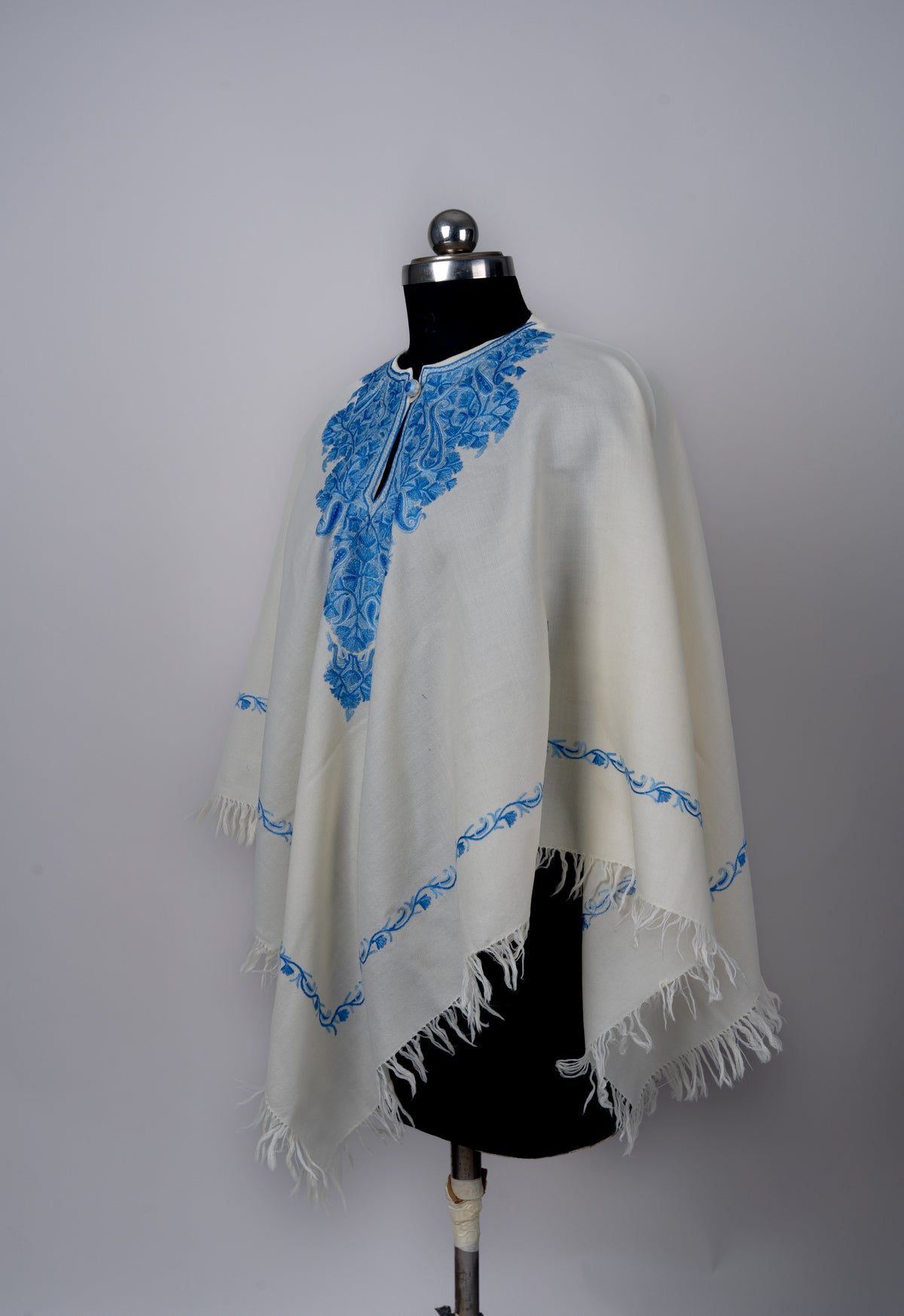 Angled front view of the ivory poncho, with blue neckline embroidery, narrow blue borders and long fringed edges.