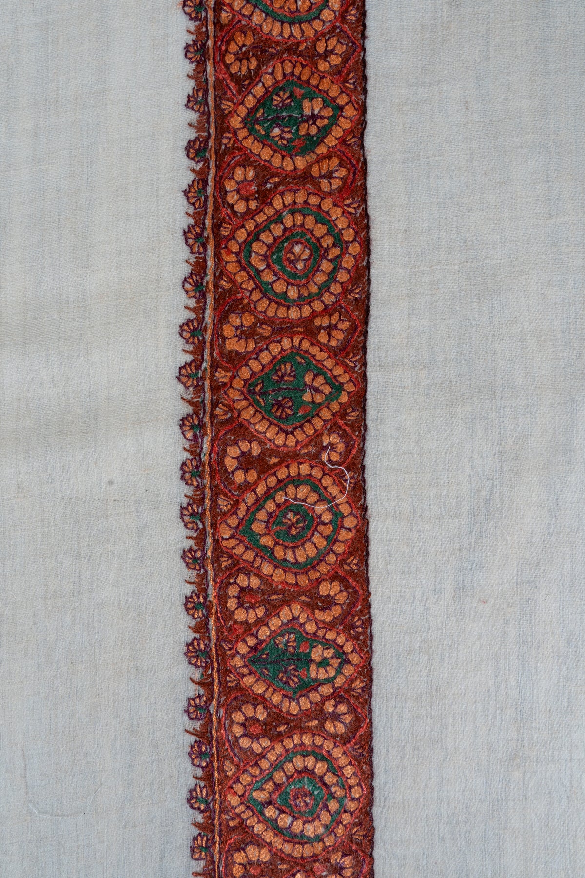 Rust-red and copper hand-embroidered border with alternating green floral medallions and fine leaf edging on ivory fabric