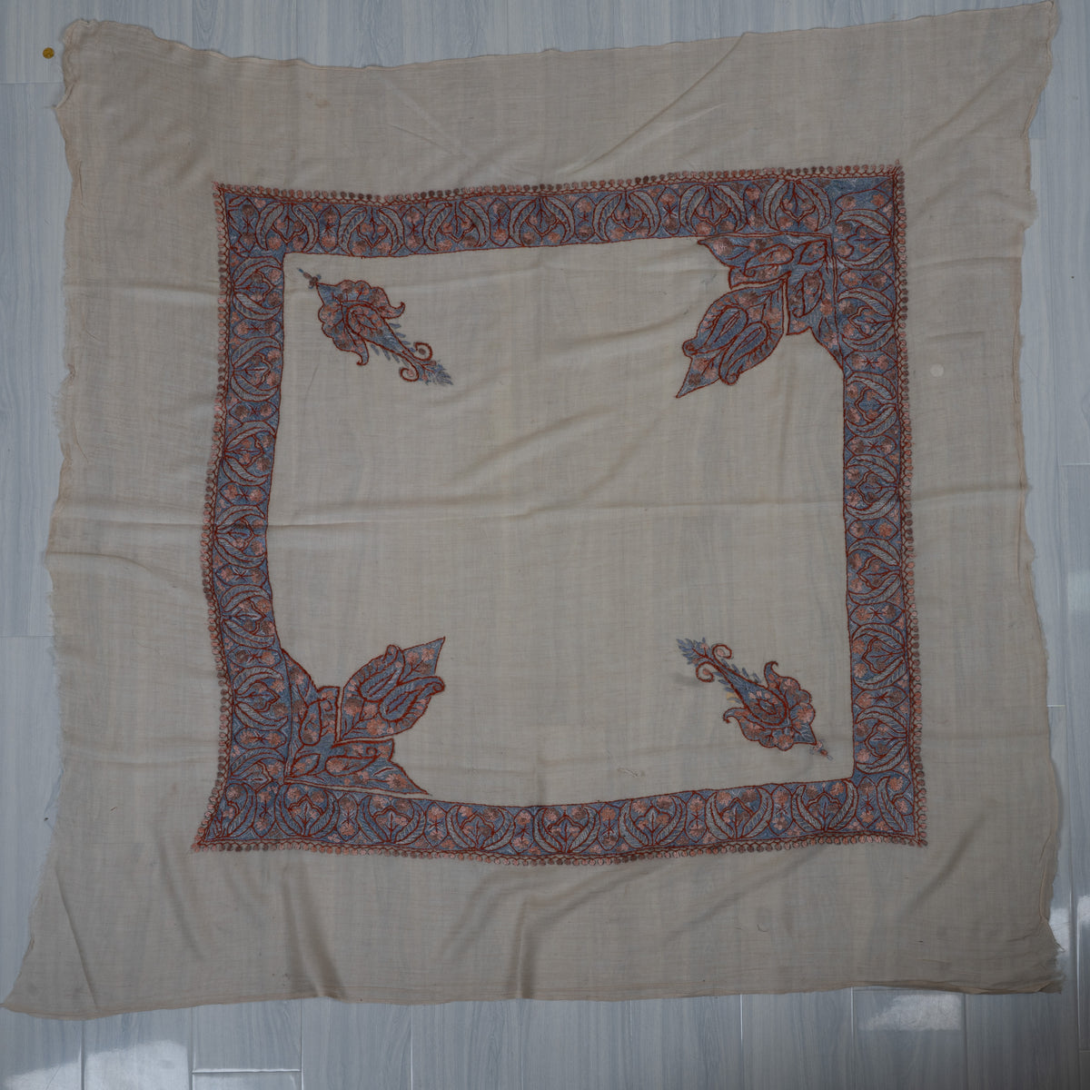 Beige square cashmere rumal laid flat with blue and dusky-rose Sozni borders, two separate paisleys and two large leafy corners.