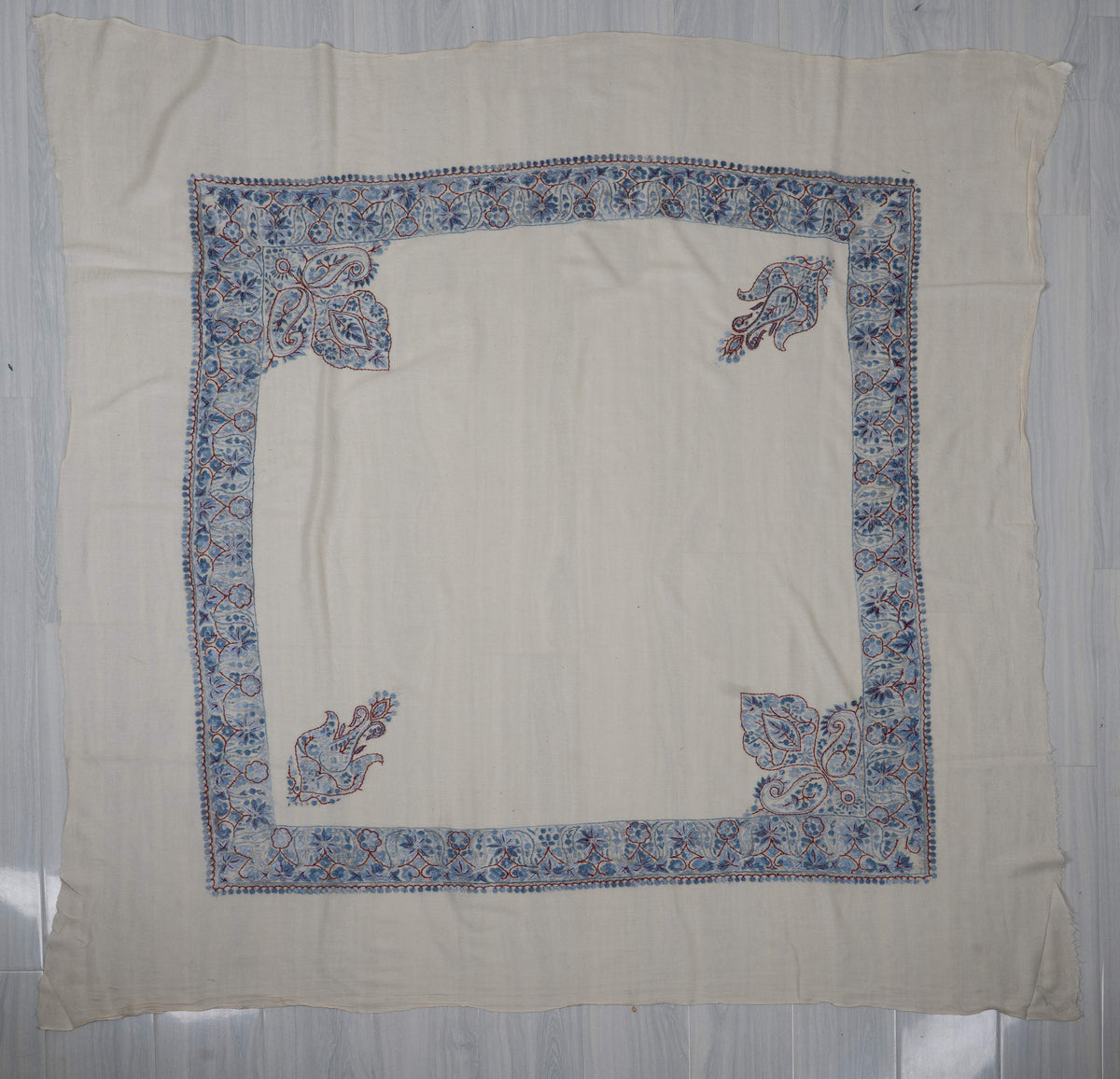 Ivory square Merino wool rumal laid flat with blue Sozni floral borders, two separate paisleys and two large leafy corners.