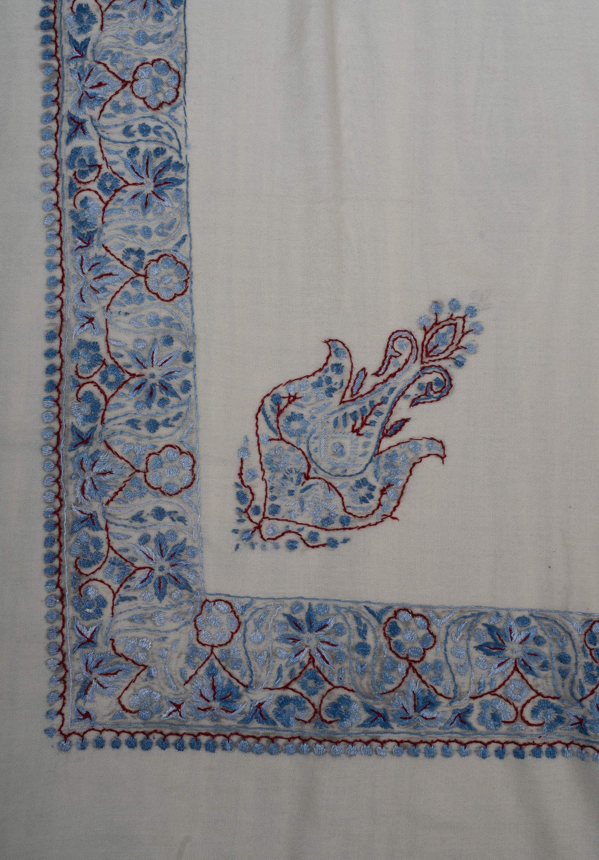 Corner of an ivory Merino wool rumal with a blue Sozni paisley and floral borders outlined in burgundy.