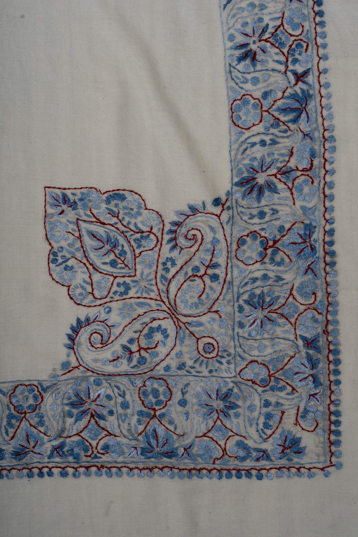 Large blue Sozni paisleys and a leaf motif rise from a floral border at one corner of an ivory Merino wool rumal.