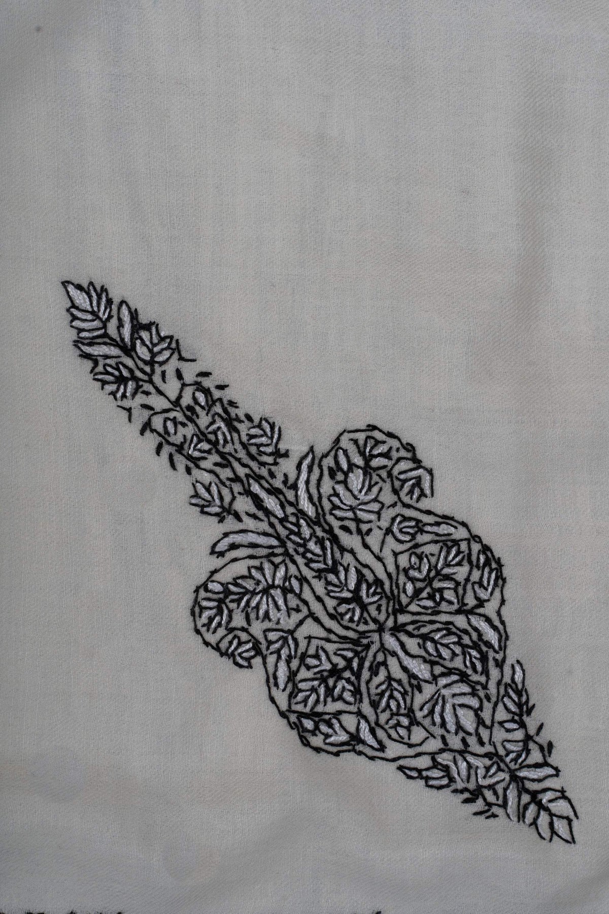 Close-up of an elongated black Sozni floral motif with curled lobes and a leafy stem on ivory rumal fabric
