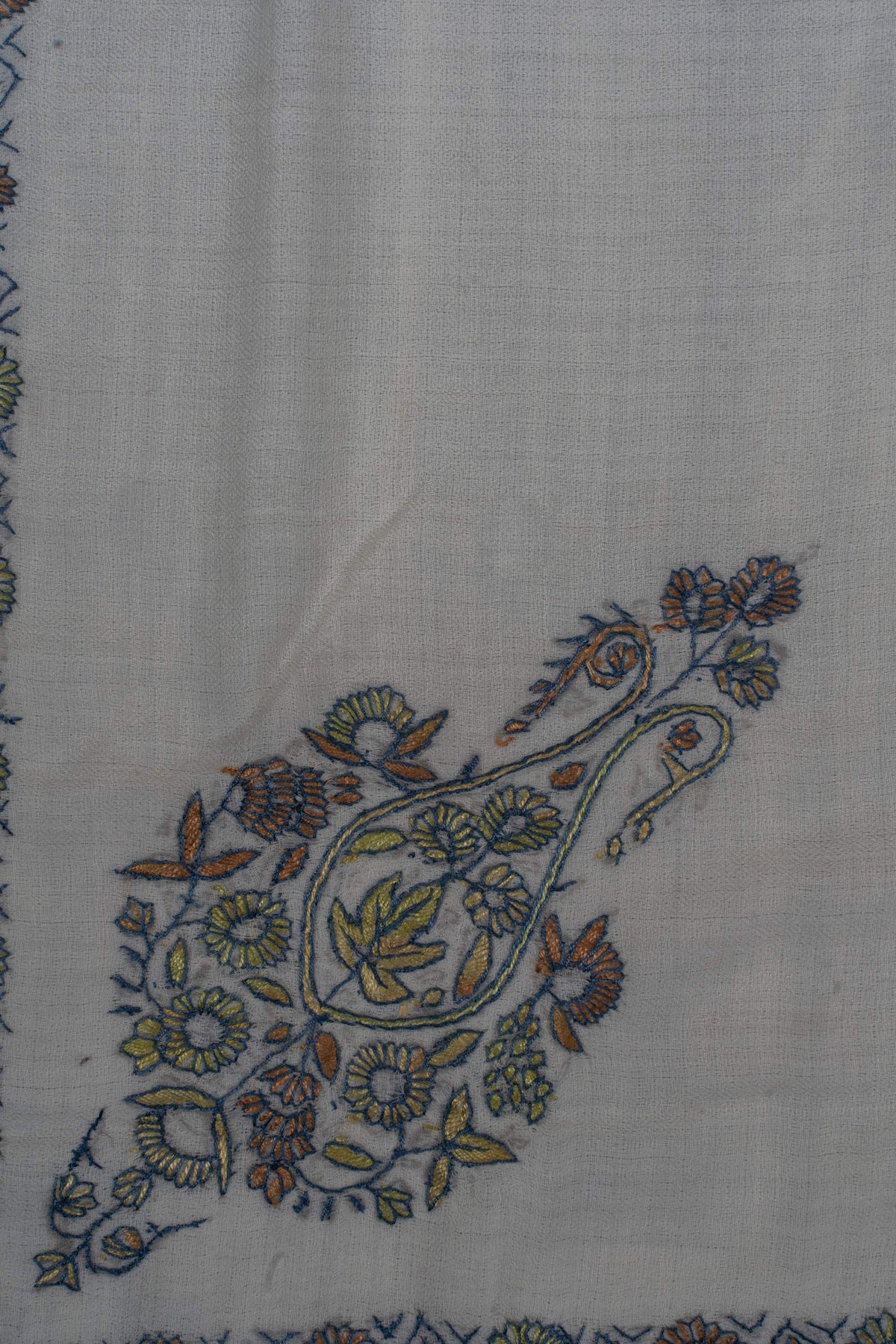 Close-up of a sage, blue and ochre Sozni paisley surrounded by small flowers and leaves on pale Merino wool.