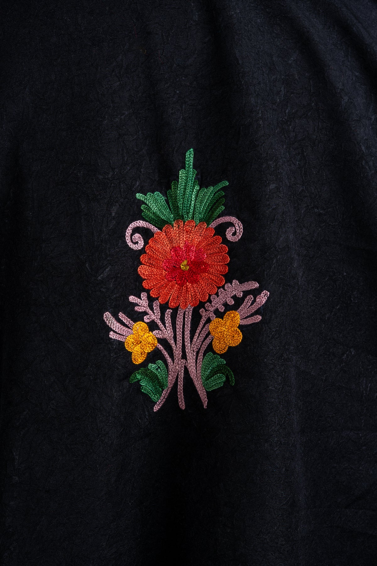 Close-up of a small Aari floral spray on black fabric, with a red-orange flower, yellow blossoms, green leaves and pink stems