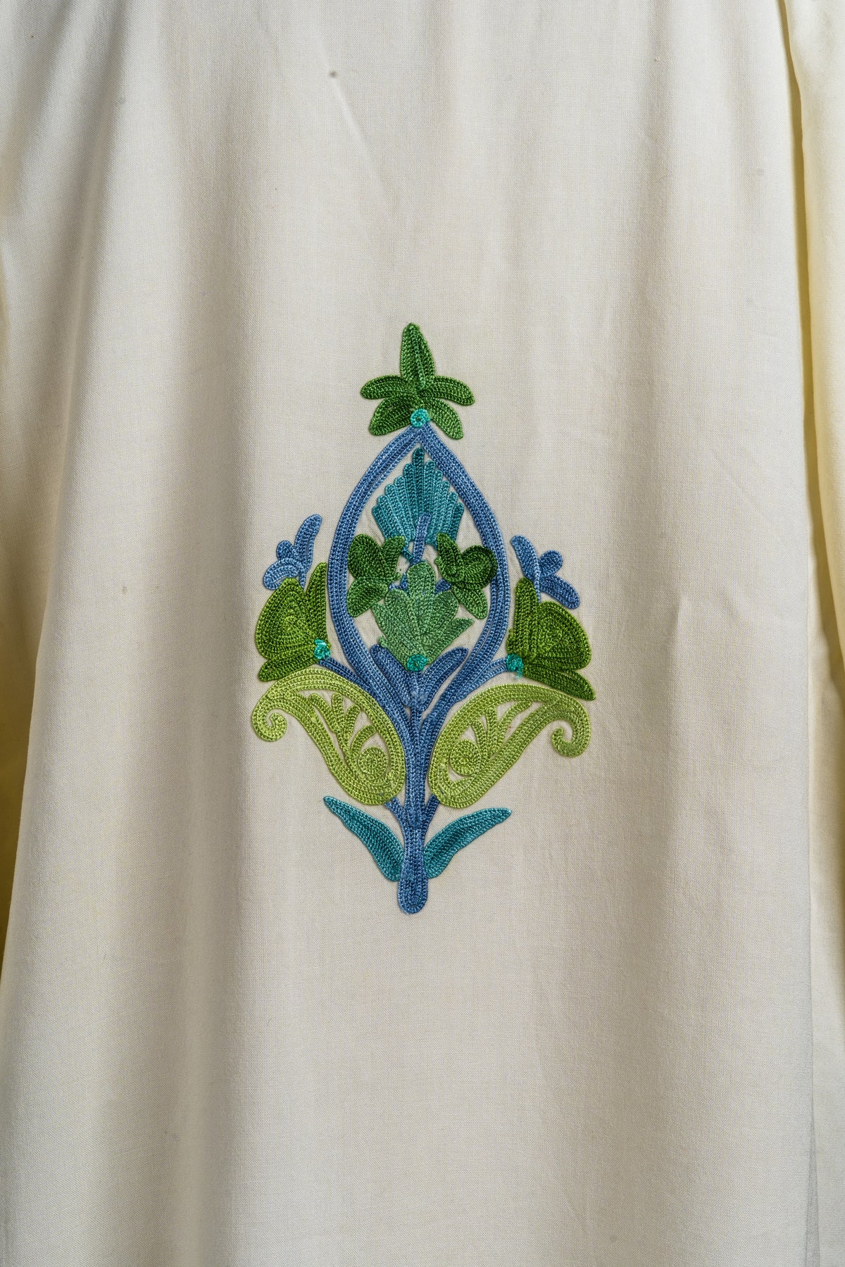 Detail of a single green and blue Aari floral motif on the ivory kaftan’s plain fabric