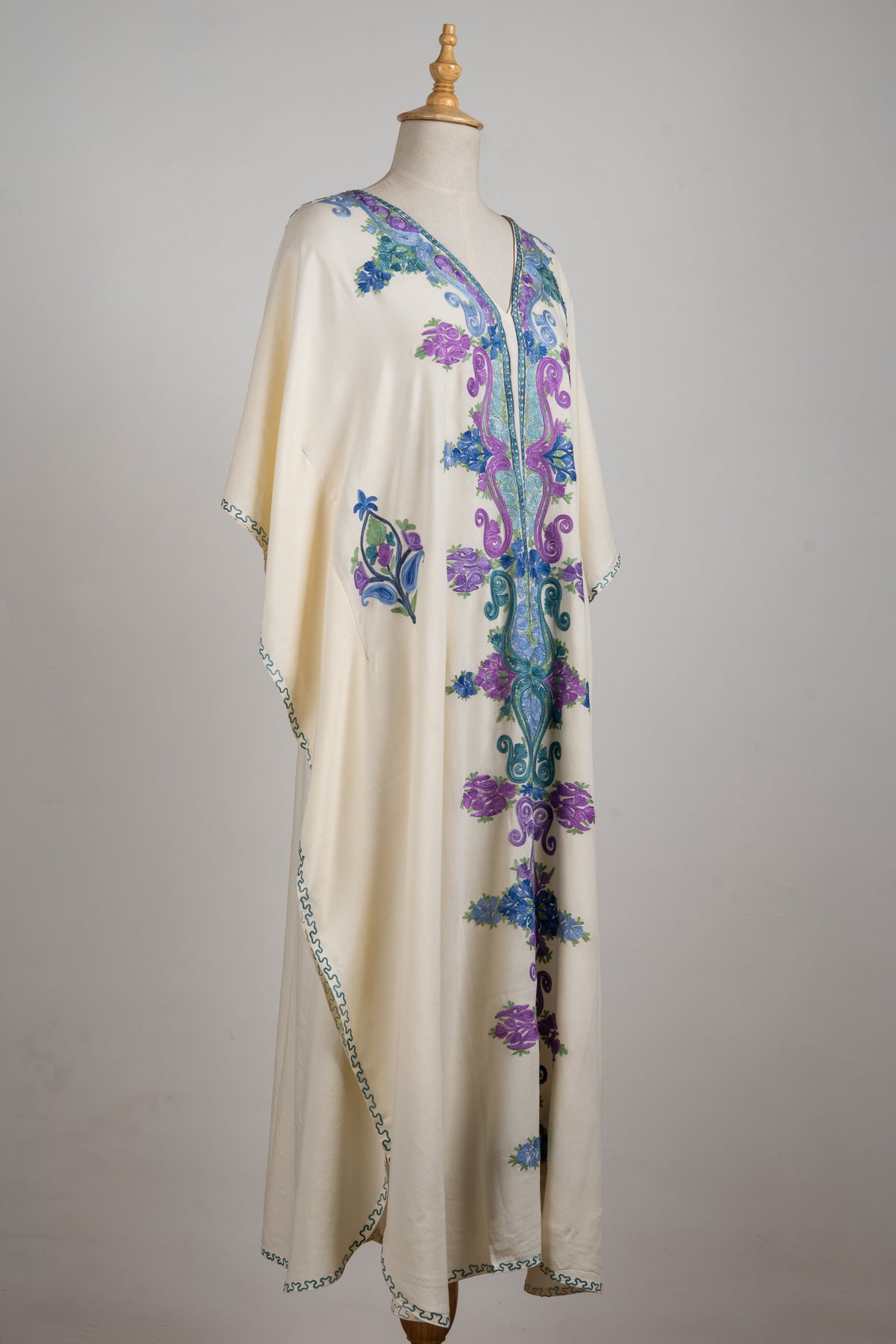 Angled full view of the ivory kaftan with flowing sleeves, a long blue and purple central design and small side motifs.