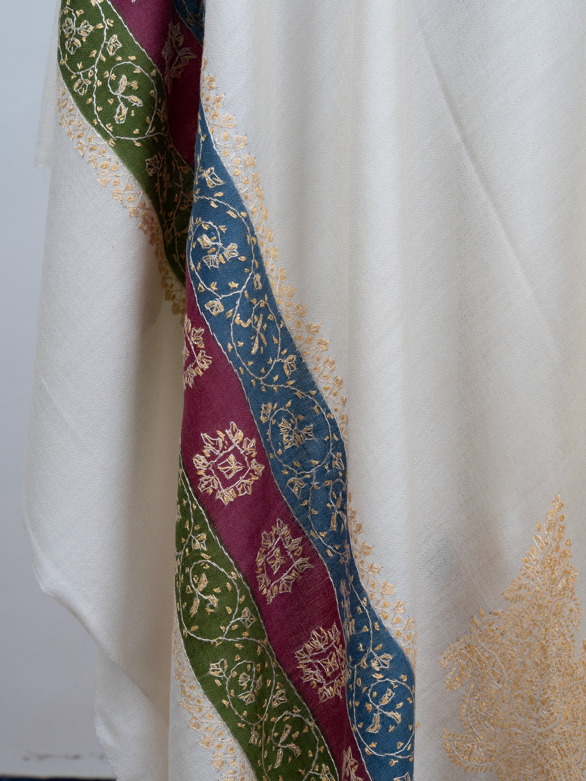 Hanging end of an ivory shawl with golden floral embroidery across blue, maroon and green bands beside a pale corner motif