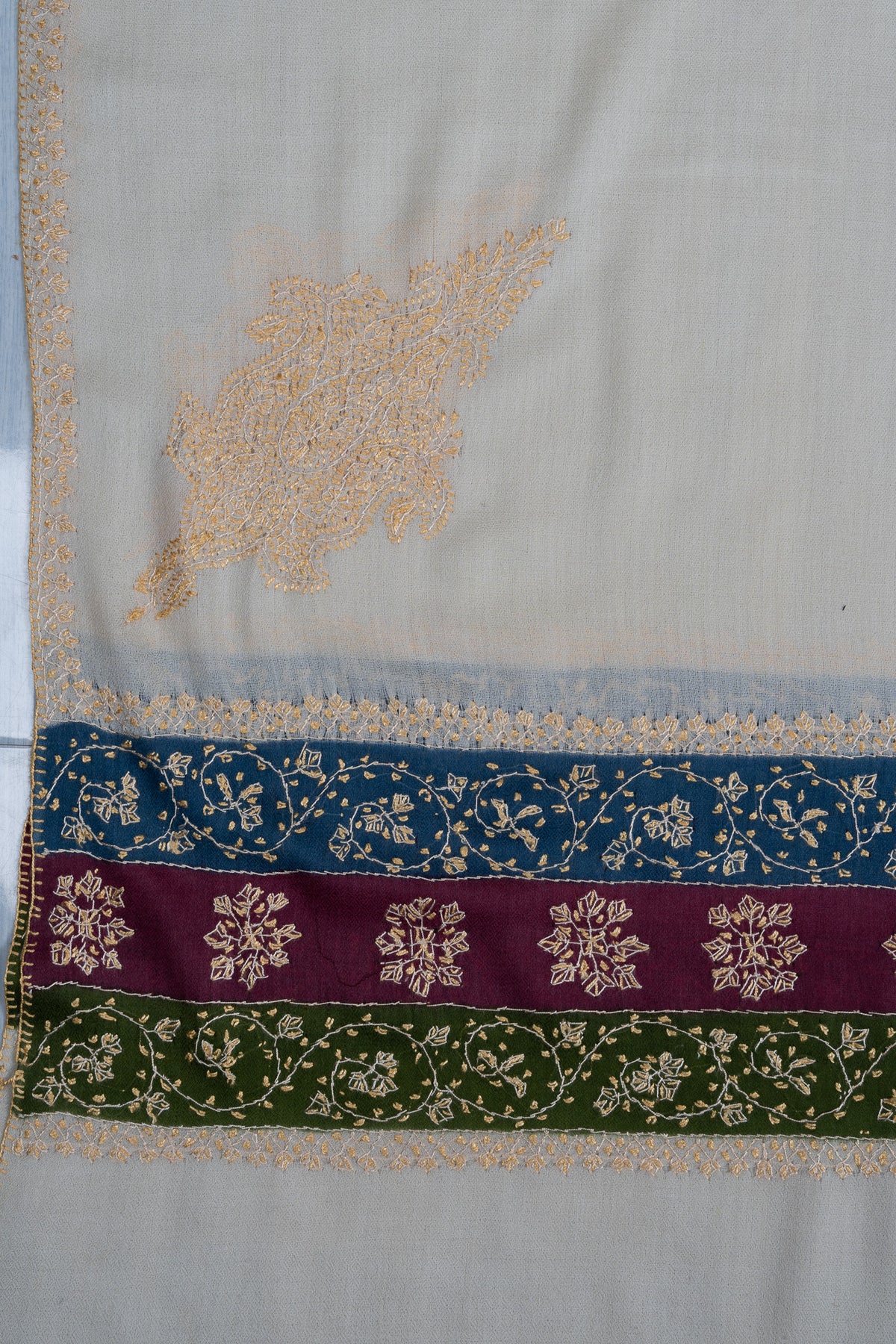 Ivory shawl corner laid flat with a pale golden motif above blue, maroon and green Sozni floral border bands