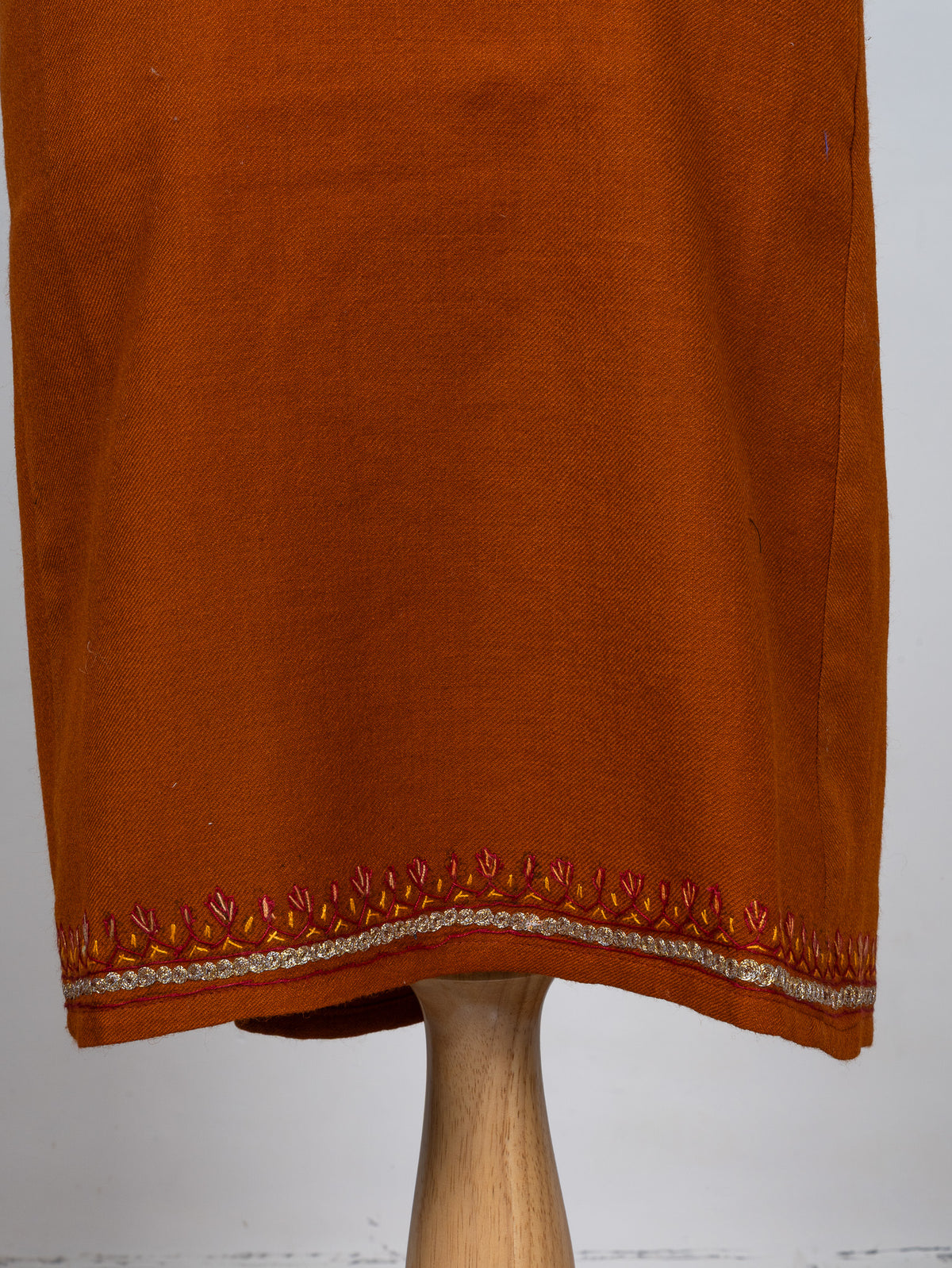 Lower front of a burnt-orange pheran with small red and gold Sozni leaves above a pale embroidered hem border