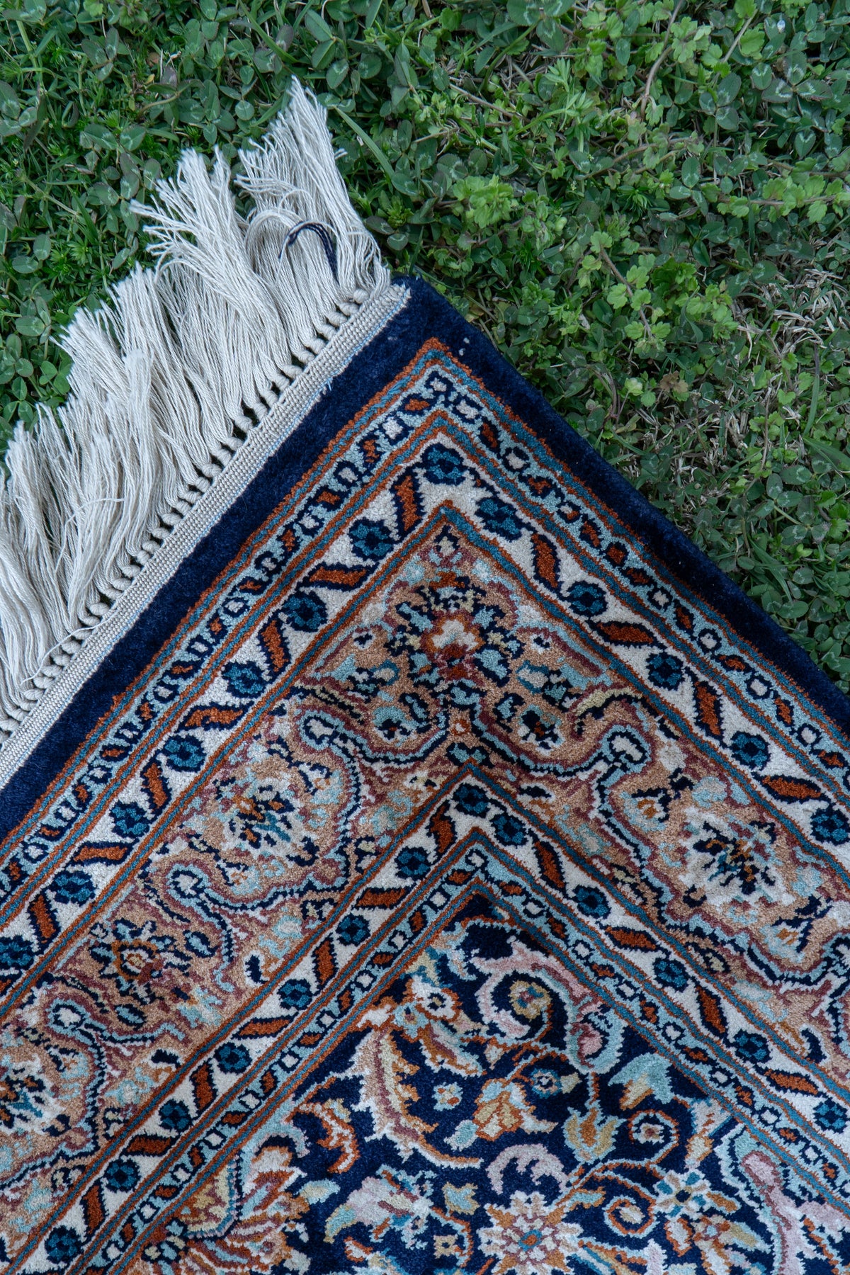 Corner of the carpet showing pale floral borders and white fringe against grass