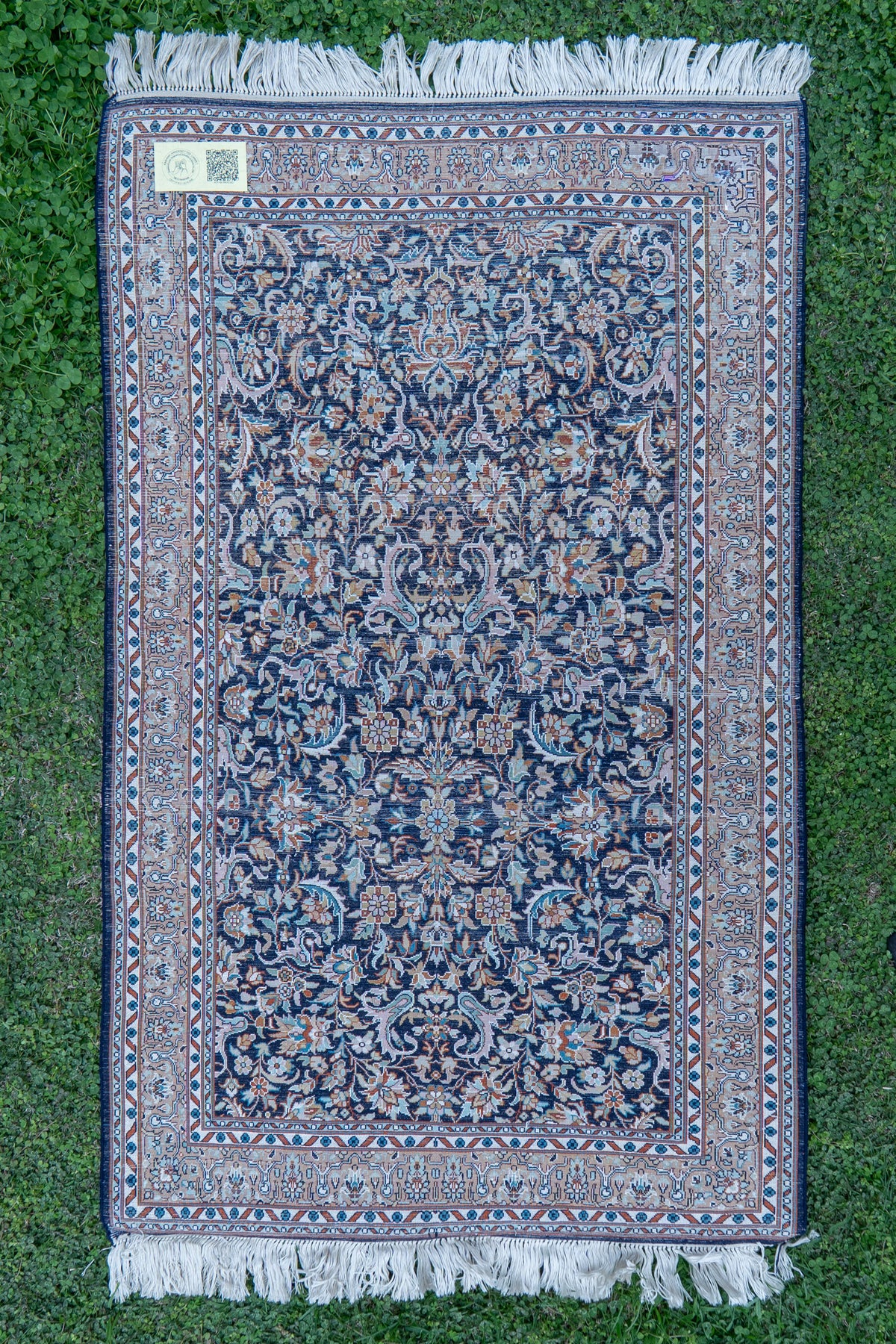 Reverse of the floral carpet laid flat on grass, showing the woven pattern, corner label and white fringe