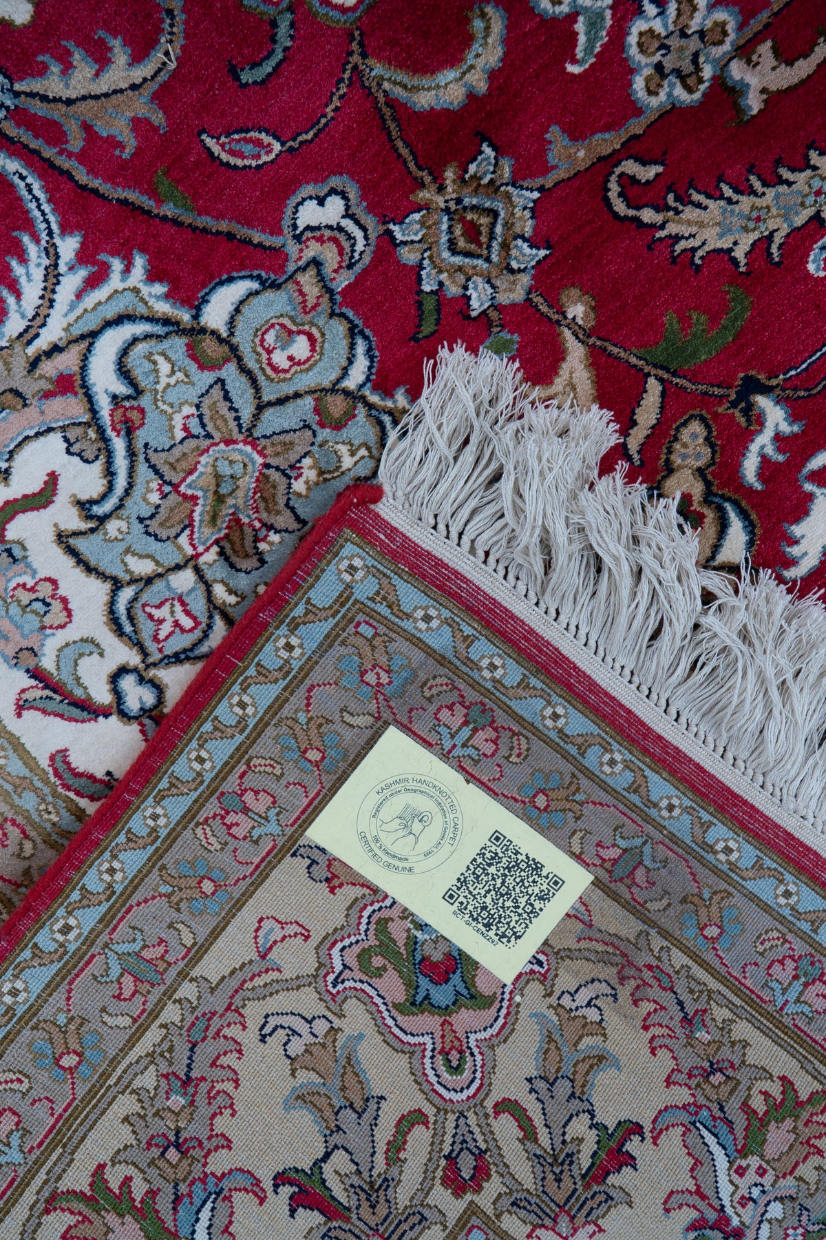 Folded corner of a crimson floral carpet showing its pale patterned underside, QR-code label and white fringe