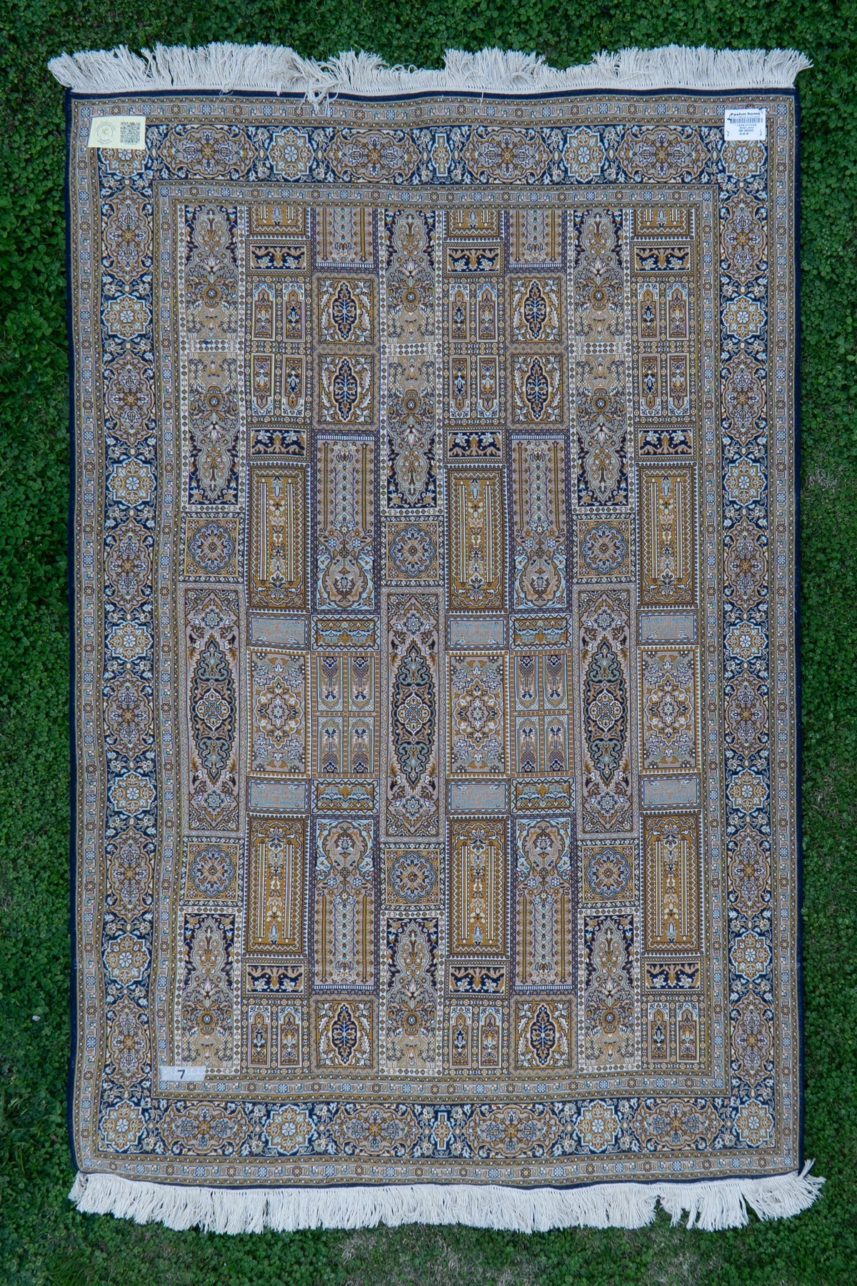 Reverse of the panel-patterned carpet laid flat on grass, with corner labels and white fringe