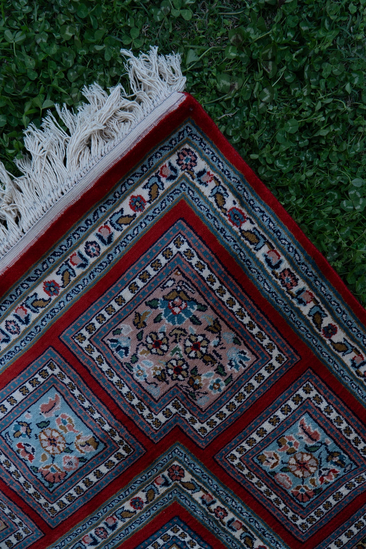 Carpet corner on grass, showing crimson-framed floral squares and white fringe