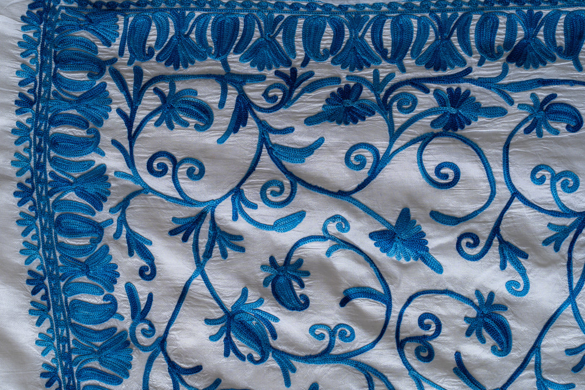 Close-up of a blue Aari border corner with repeated leaf shapes, small floral edging and curling vines on ivory fabric