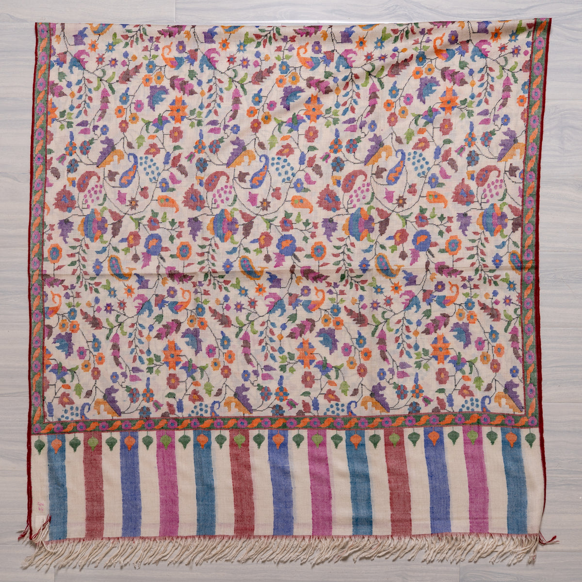 Ivory Pashmina shawl spread flat, with an all-over multicoloured floral pattern above a pink-and-blue striped fringed end.