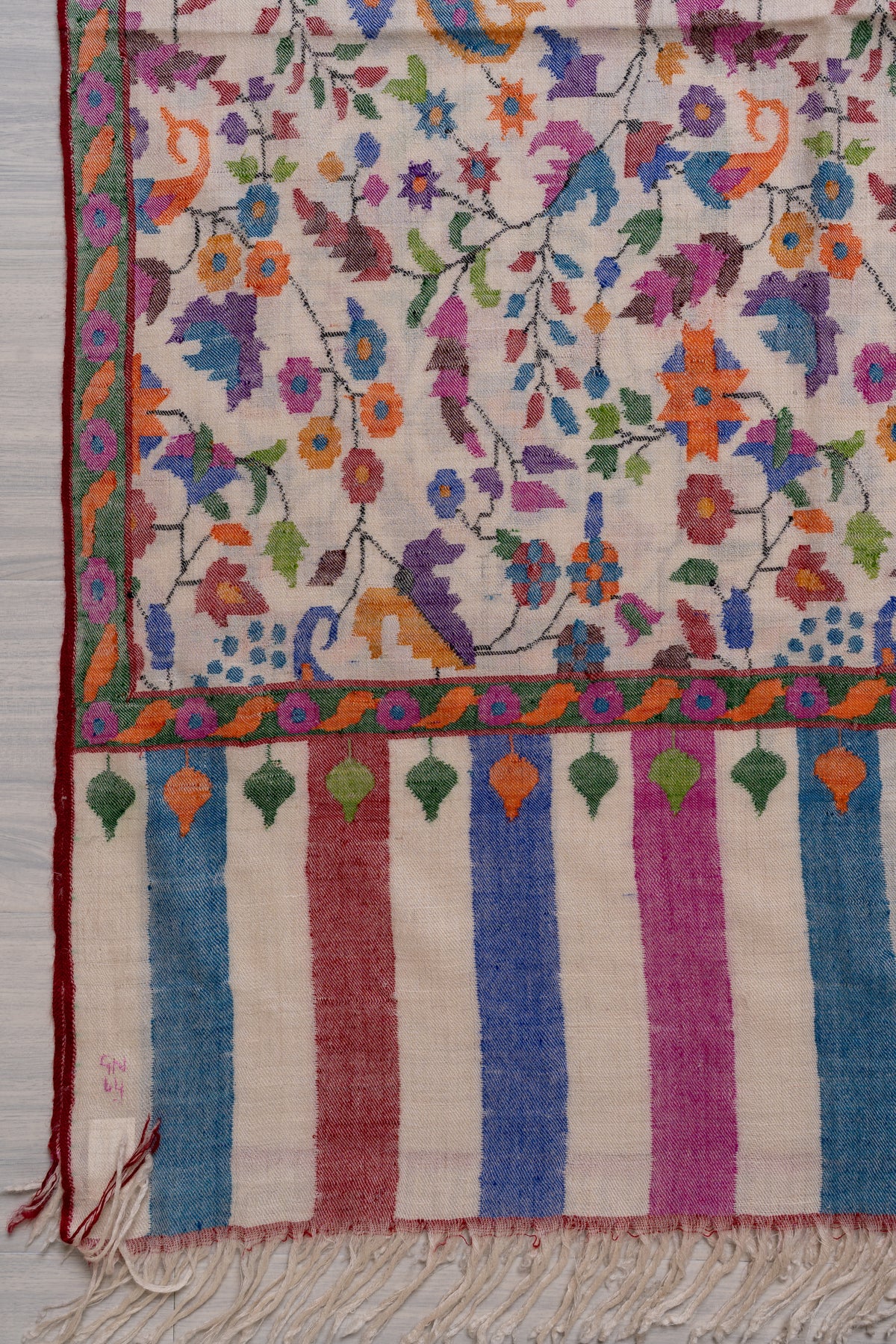 Corner of an ivory Pashmina shawl laid flat, with colourful floral vines and borders above pastel stripes and ivory fringe.