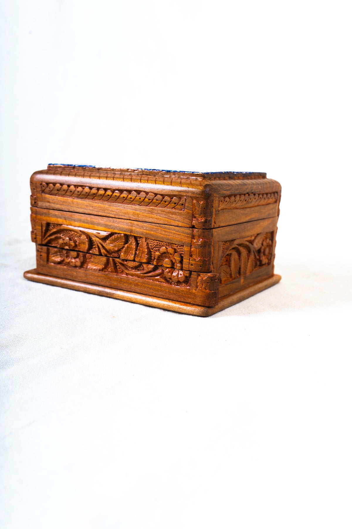 Low angled view of a closed walnut wood box with carved flowers and leaves along the sides and a raised decorative top