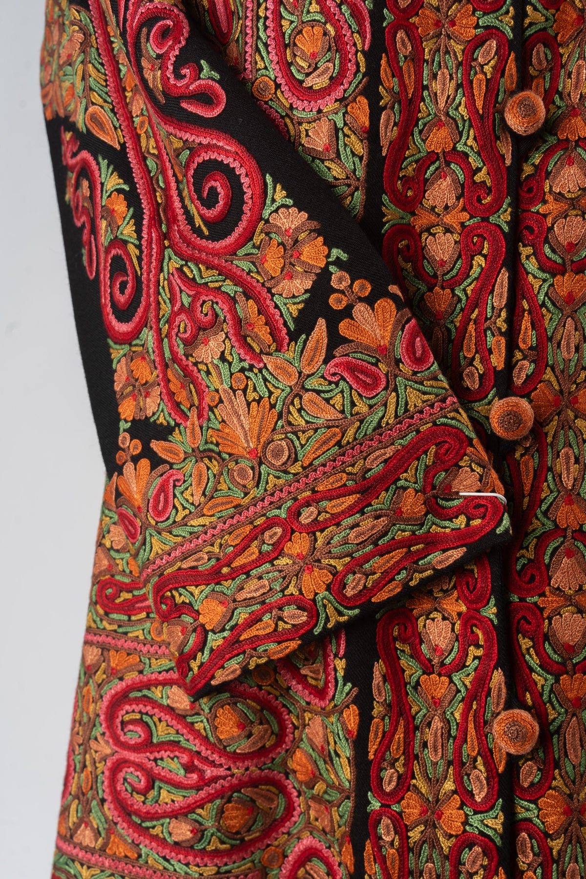 Crossed jacket cuff with red Aari scrollwork and orange flowers over the densely embroidered front of a black Merino wool jacket