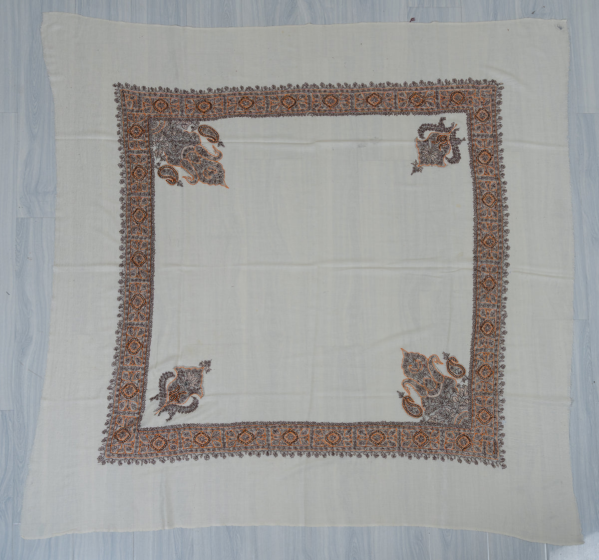 Ivory square Merino wool rumal laid flat, with a charcoal and ochre embroidered border and four ornate corner motifs