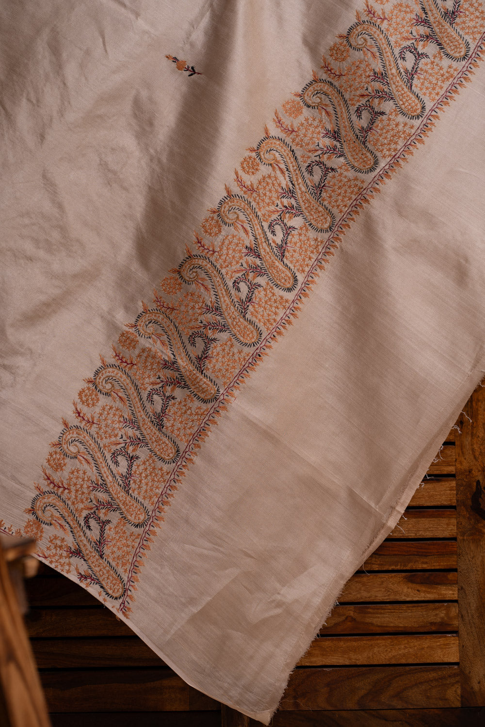 Draped corner of the ivory tabby silk saree, showing a narrow orange-and-blue Sozni paisley border and small scattered sprigs.