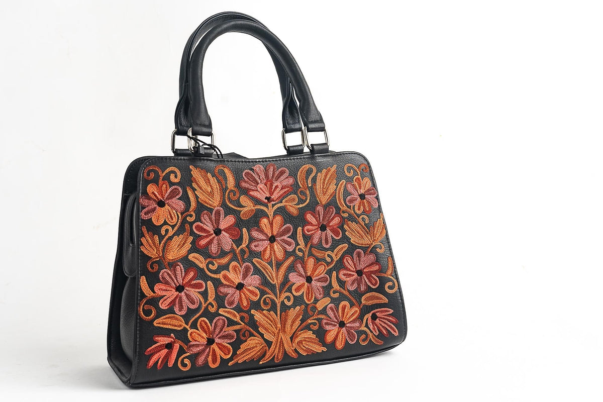 Angled front view of a black two-handle handbag with coral-pink and orange floral embroidery and silver-tone handle fittings