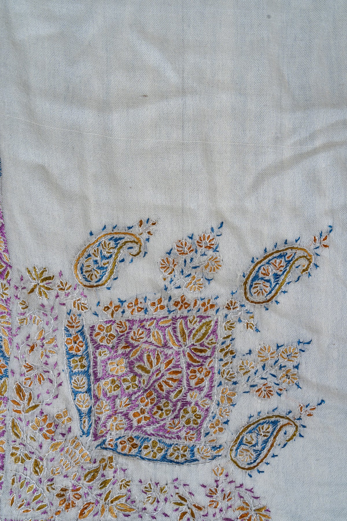Close-up of a purple Sozni floral medallion surrounded by small blue paisleys and gold leafy sprigs on an ivory rumal