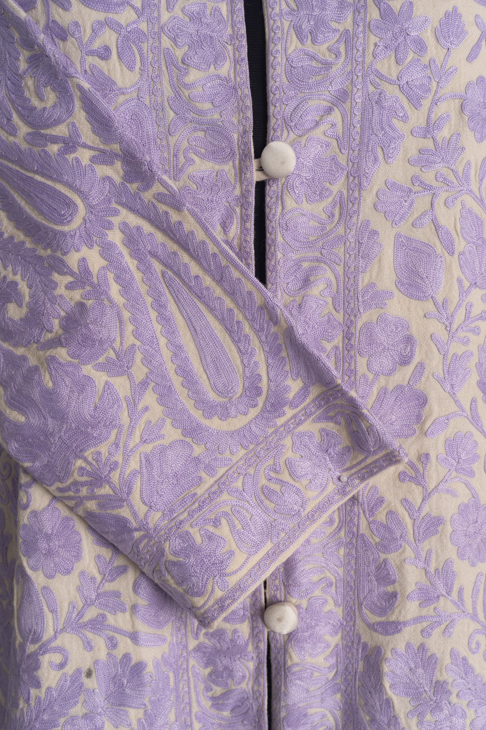 Close-up of a lilac paisley on the cream jacket’s crossed cuff beside embroidered front borders and covered buttons