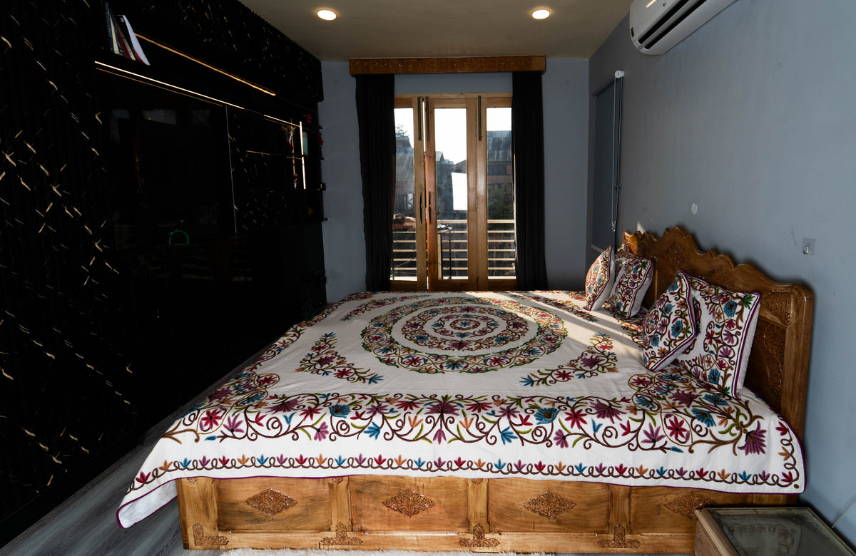 Room view of a wooden bed dressed in the cream crewel set, with a circular floral centre and matching embroidered pillows.