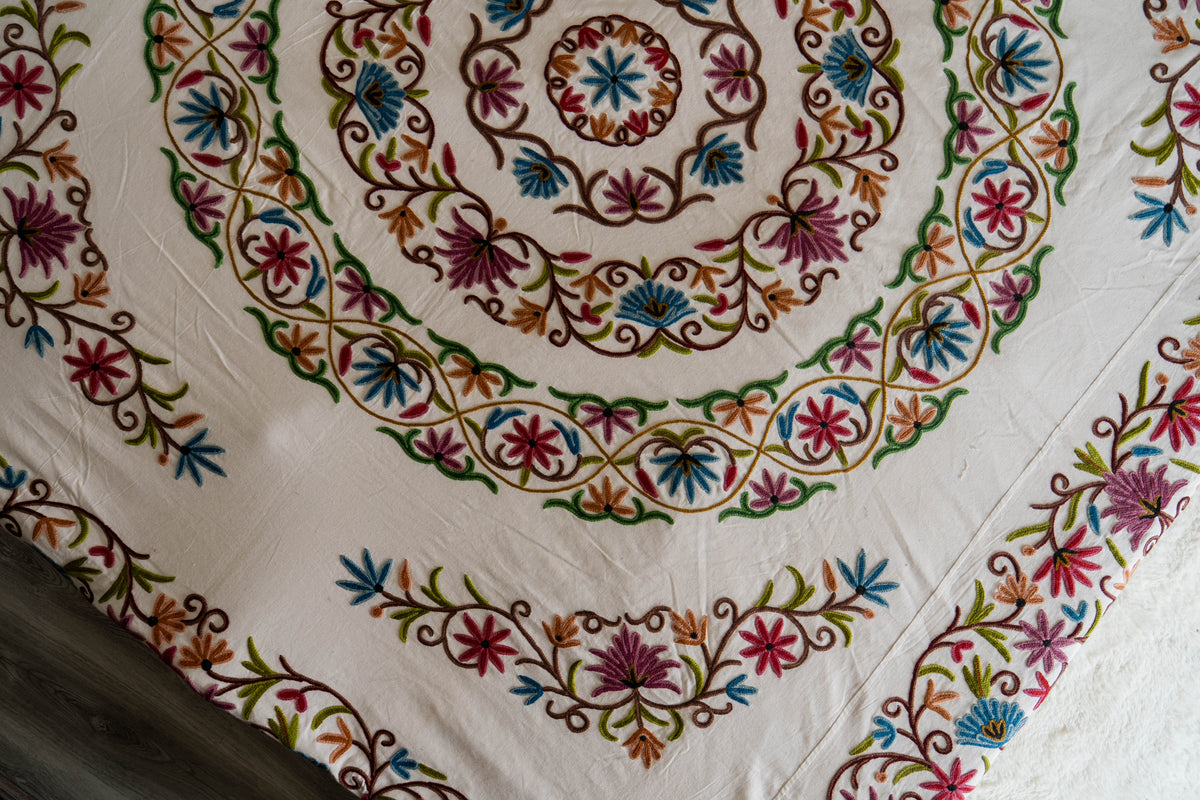 Close view of the cream cotton bed cover with concentric crewel floral rings and colourful embroidered corner sprays.
