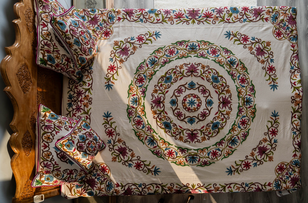 Overhead view of the cream bed cover with a large circular floral centre, embroidered borders and matching pillows and cushions.