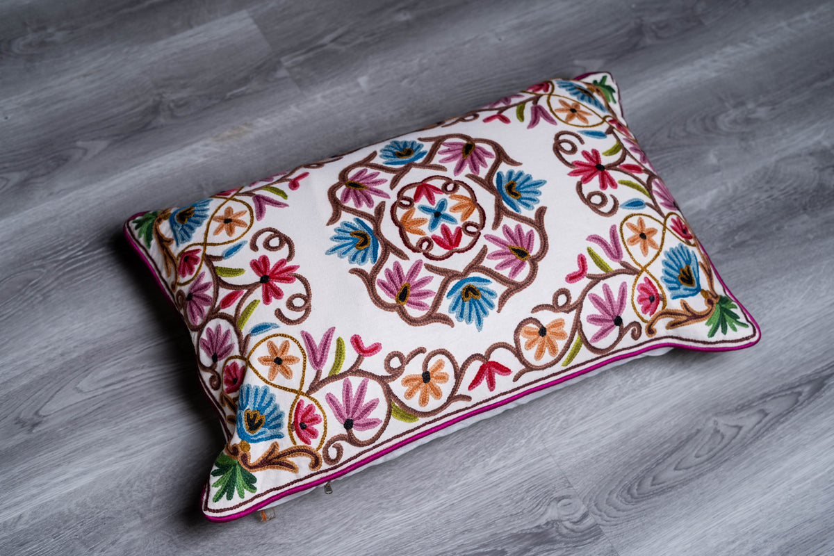 Rectangular cream pillow with colourful crewel flowers, curling vines and burgundy piping, placed on a grey floor.