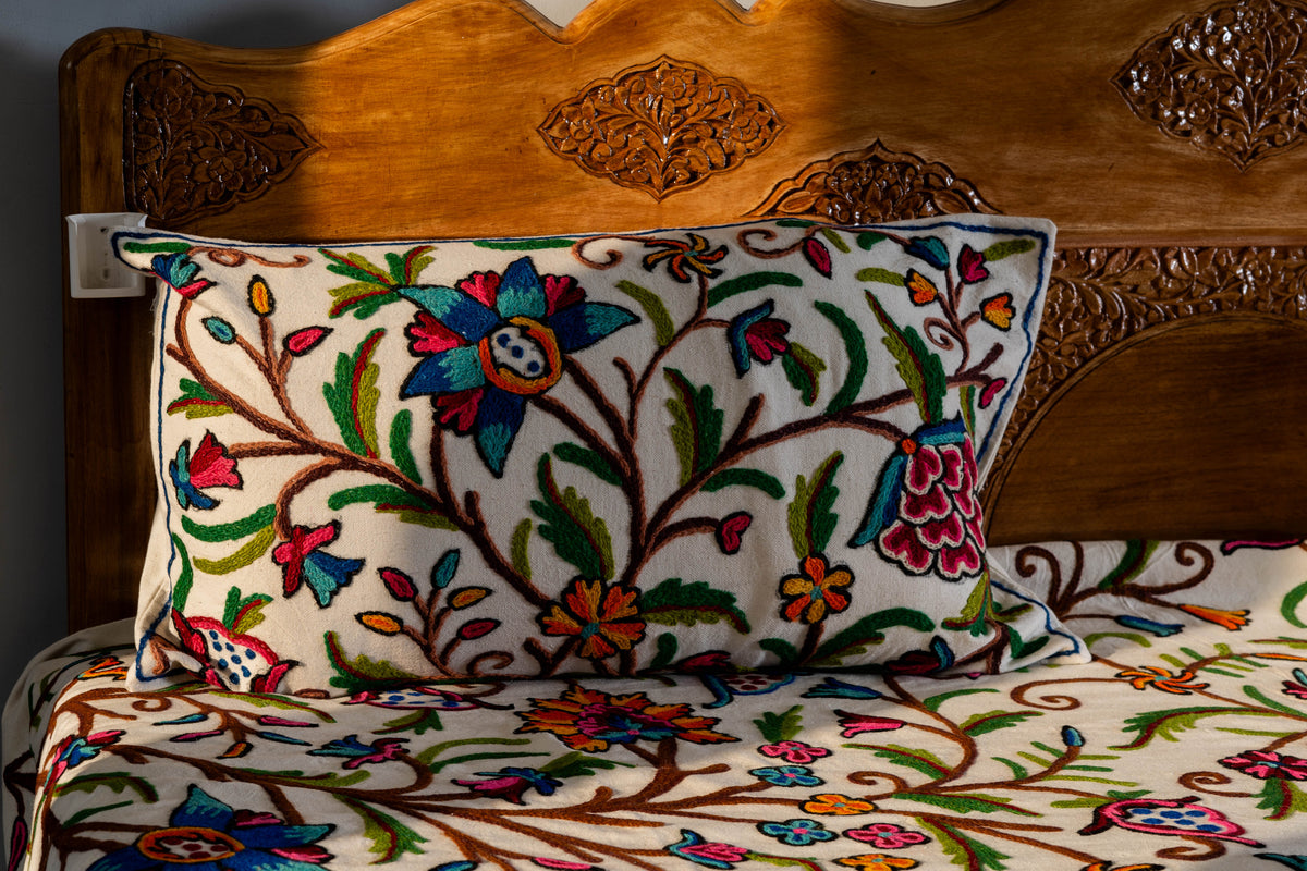 Rectangular cream pillow with bright floral and vine embroidery resting on the matching bed cover against a wooden headboard.