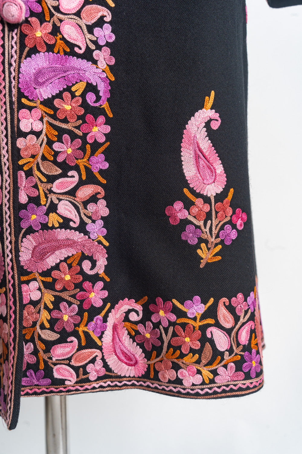 Lower front corner of the black coat with pink paisley motifs above a colourful embroidered floral hem