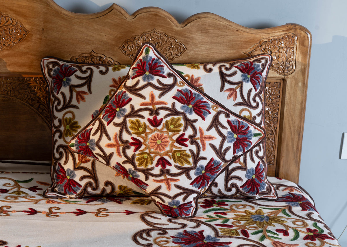 Cream embroidered cushion set against a matching rectangular pillow, with red, blue and mustard flowers before a carved wooden headboard