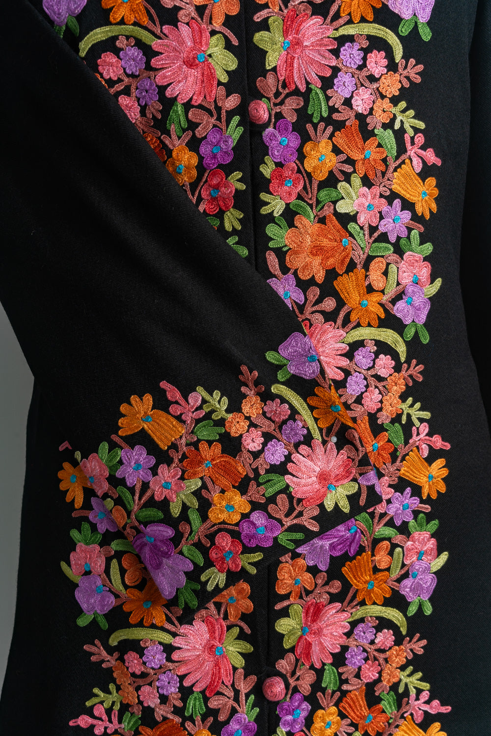 Close-up of bright Aari floral embroidery on the black jacket’s folded cuff and buttoned front panel.