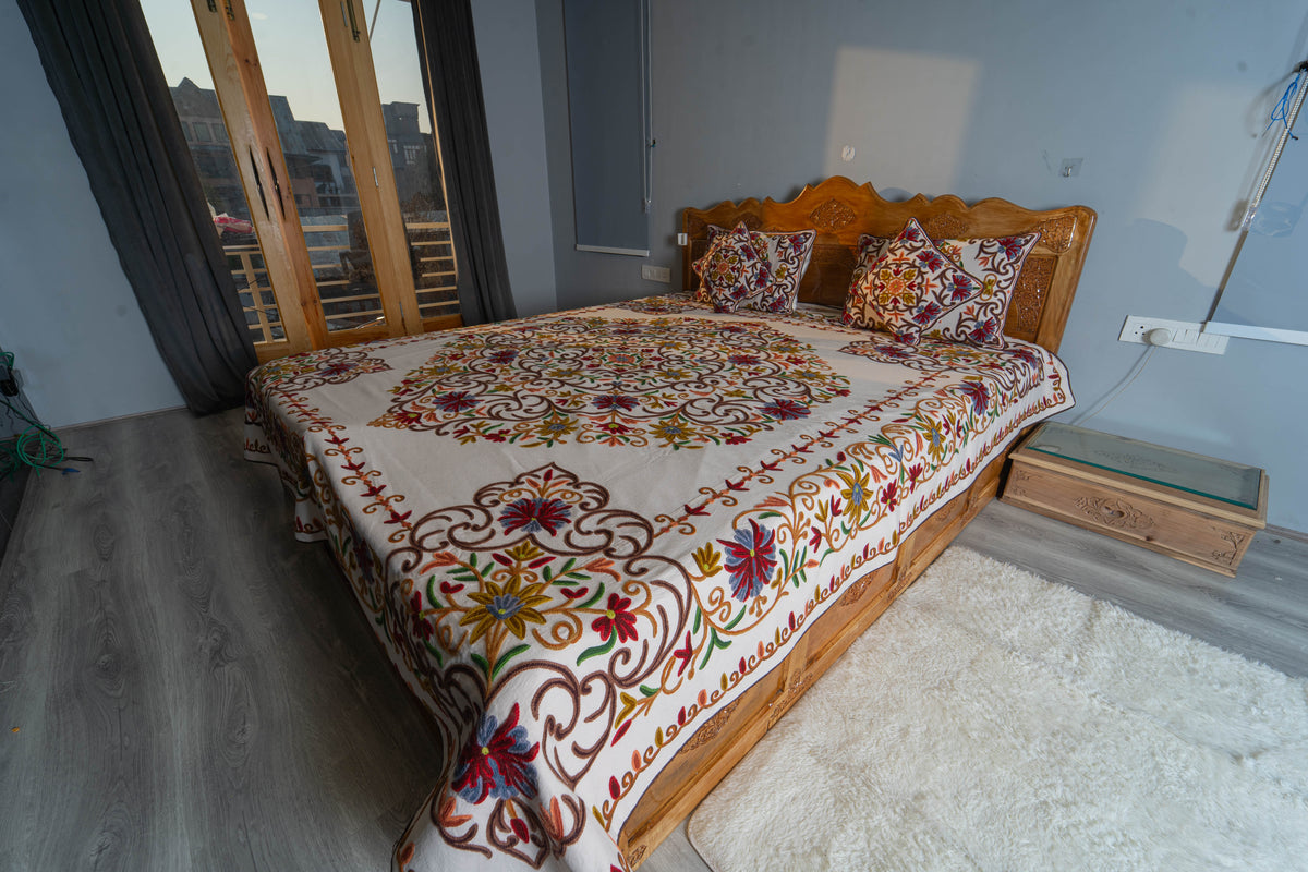 Angled bedroom view of the cream Crewel bed cover set, with a large floral medallion and matching cushions on a wooden bed