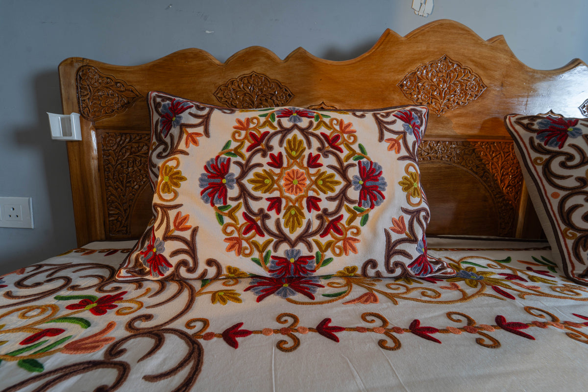 Square cream cushion with a circular floral medallion, displayed against matching embroidered pillows on the bed
