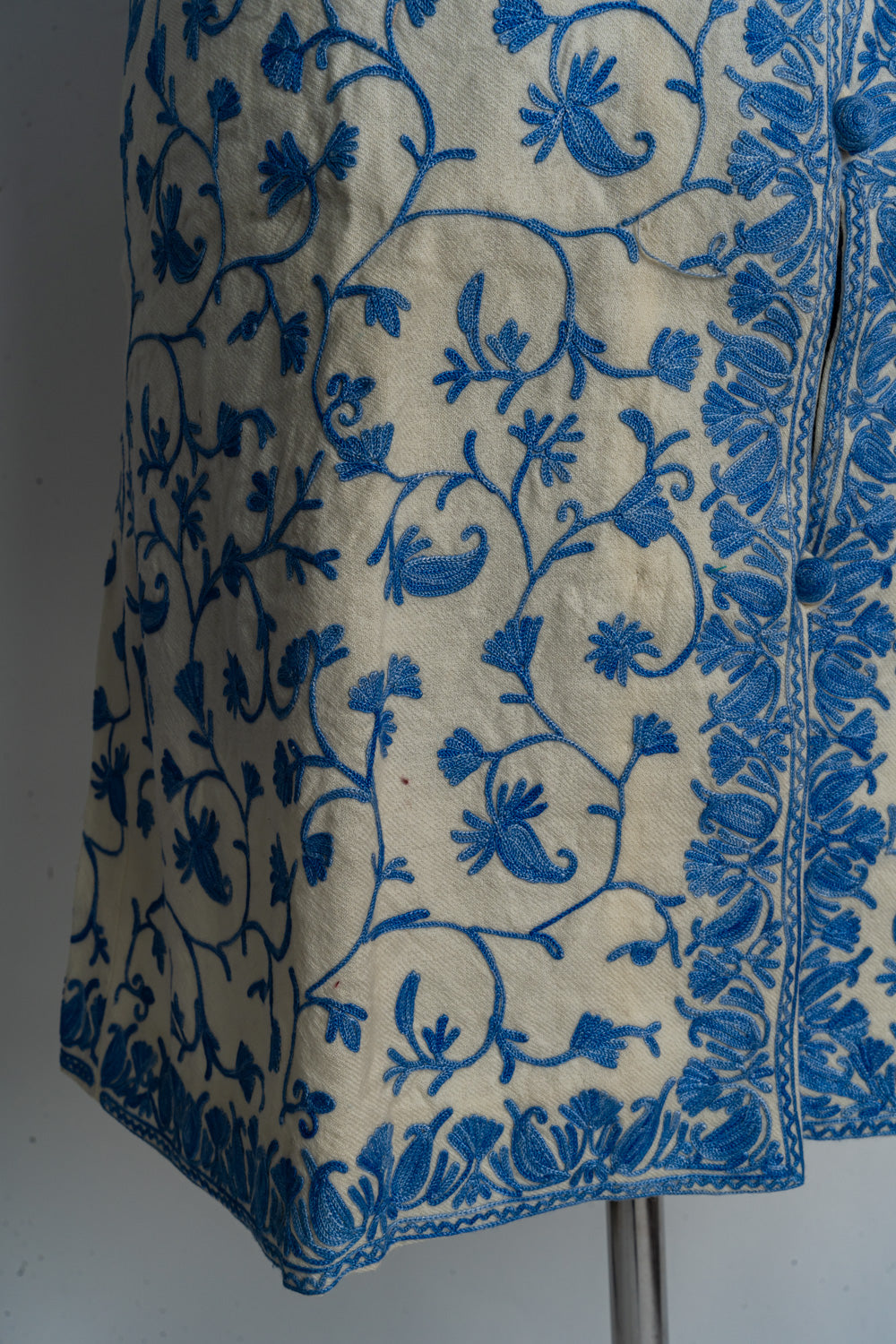 Lower front corner of the cream jacket with blue floral vines and an embroidered border around the opening and hem.