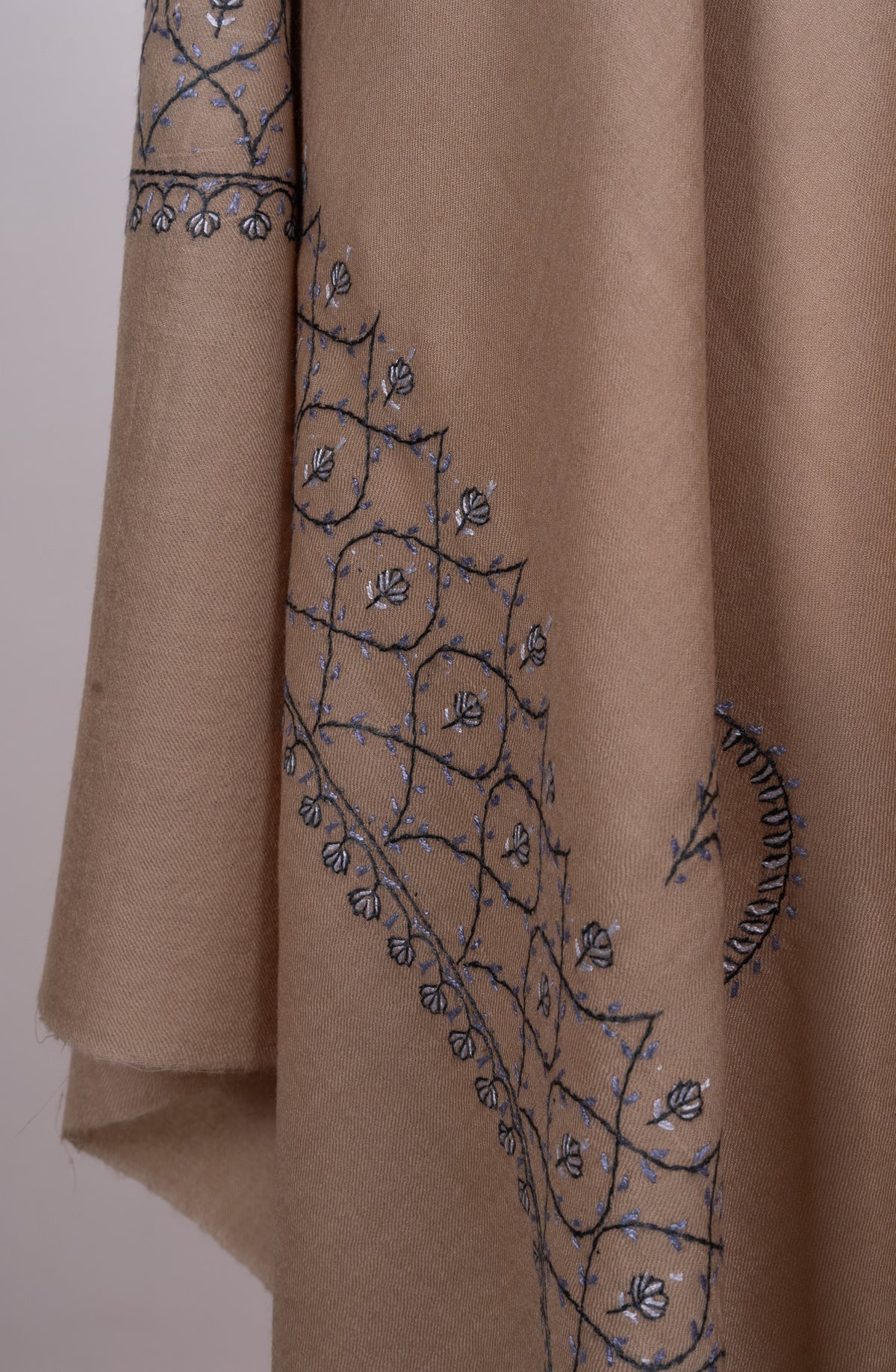 Desert-beige shawl hanging in folds, with blue-grey lattice embroidery and small flowers along the draped border