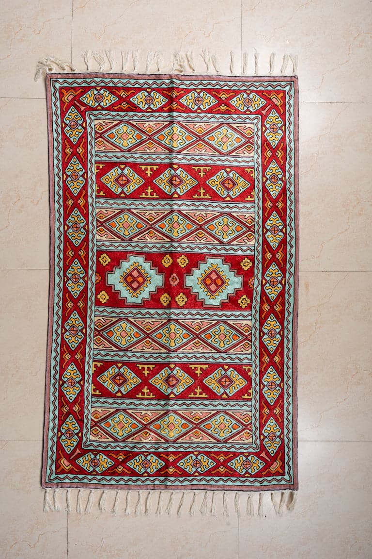 Overhead view of a red geometric rug with turquoise stepped medallions, patterned horizontal bands and white fringe at both ends