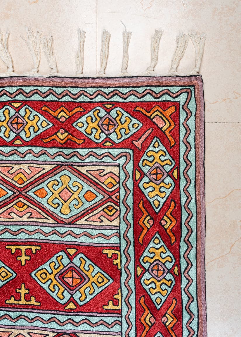 Corner of the red geometric rug showing turquoise and yellow diamond motifs, pink edging and knotted white fringe