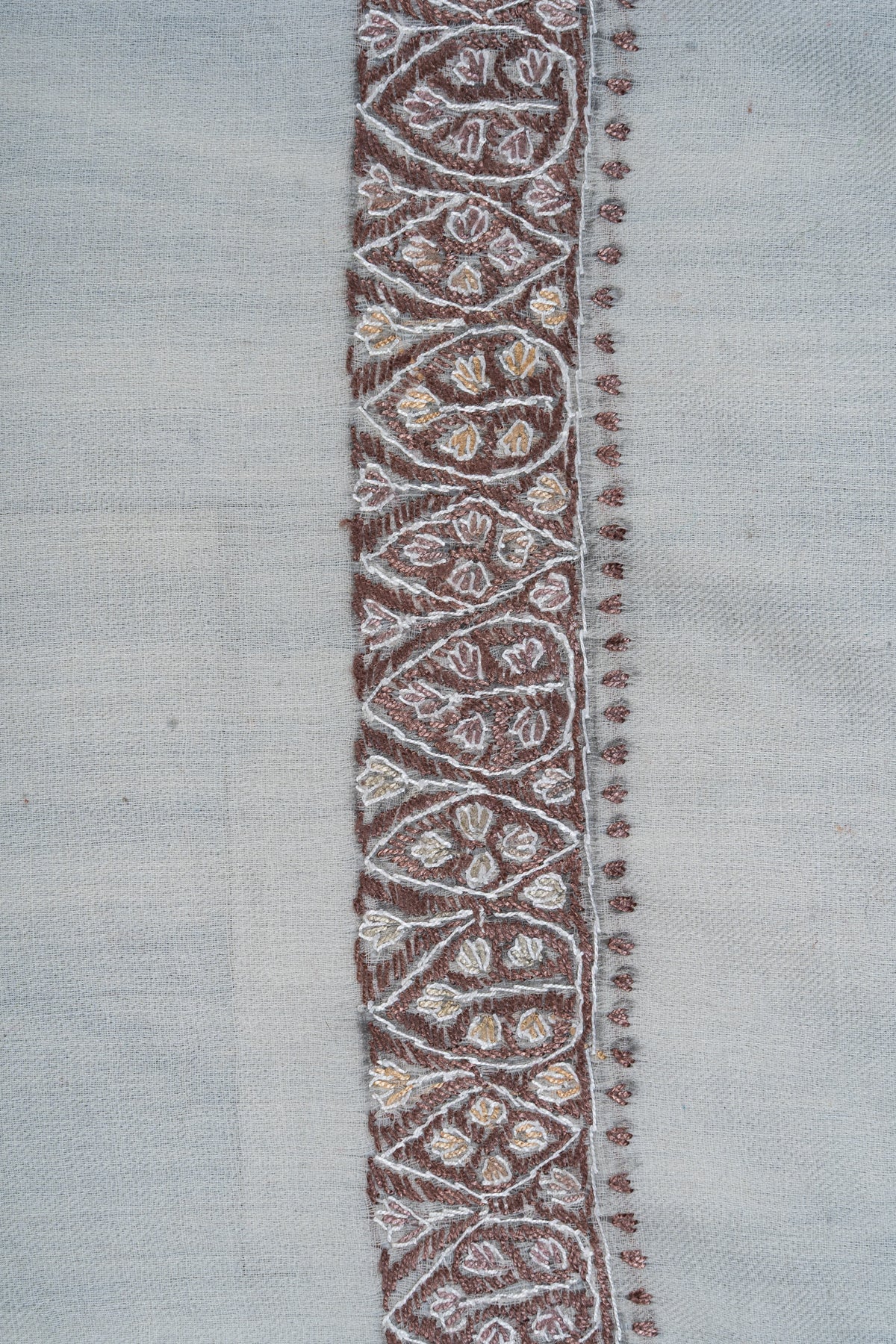 Close-up of a mocha rumal border filled with ivory-outlined leaves and geometric shapes beside tiny stitched edging
