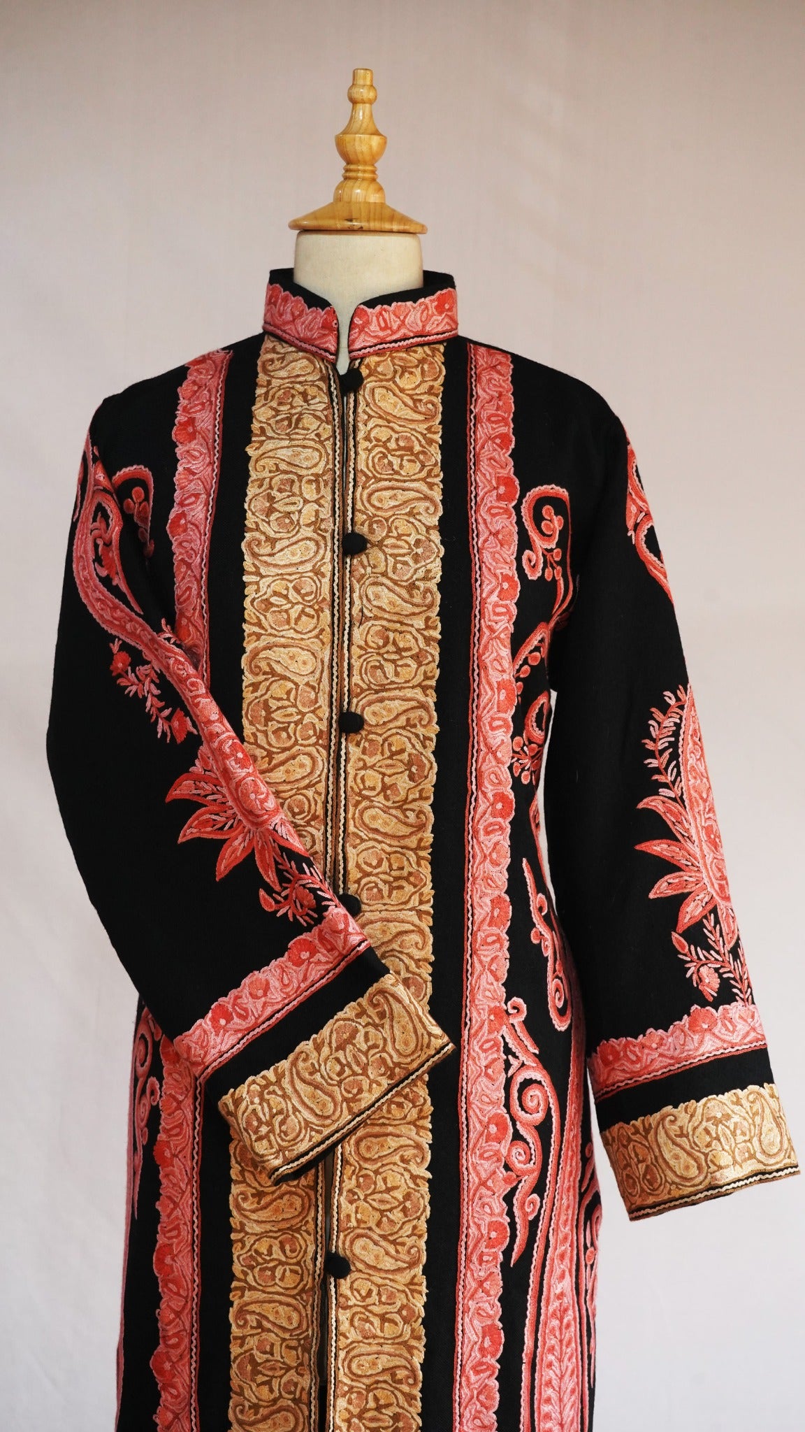 Upper front of the black jacket on a mannequin, with a coral-pink stand collar, golden paisley panels and a crossed sleeve